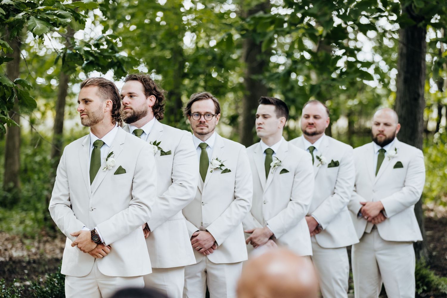 Groomsmen in crisp white suits with green boutonnieres stand in line during an outdoor wedding ceremony.