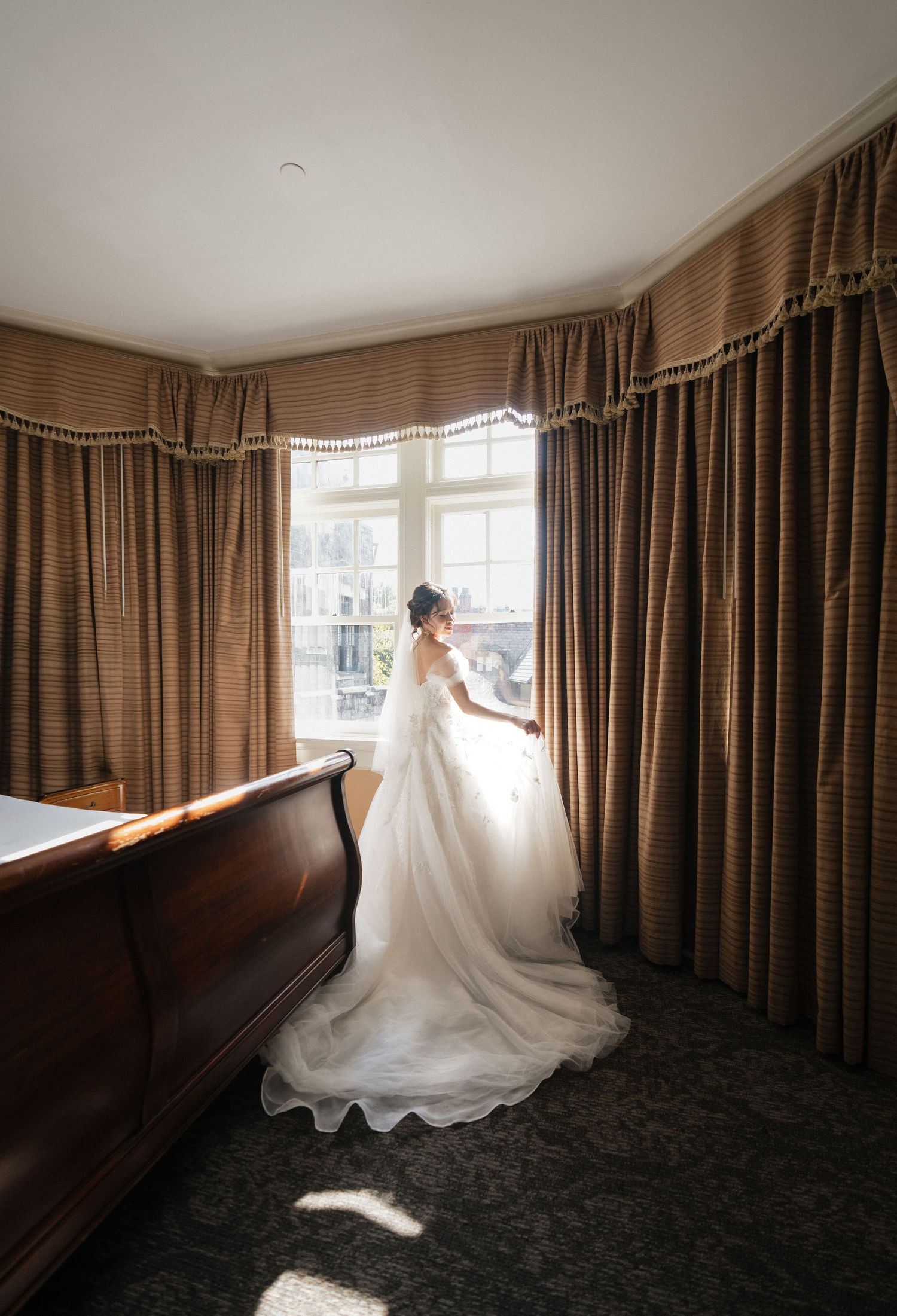 Bride in a white ball gown standing by a sunlit window with brown curtains in an elegant hotel room.