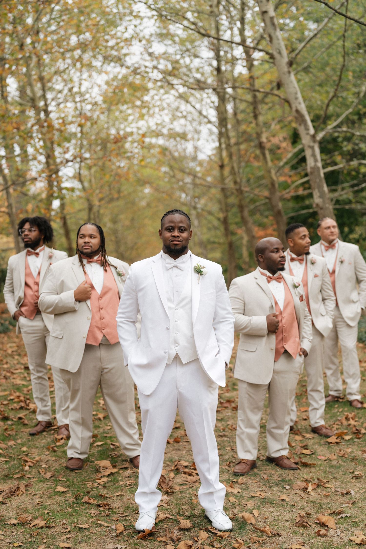 Groomsmen in white and cream suits pose playfully for photos in a wooded autumn setting.