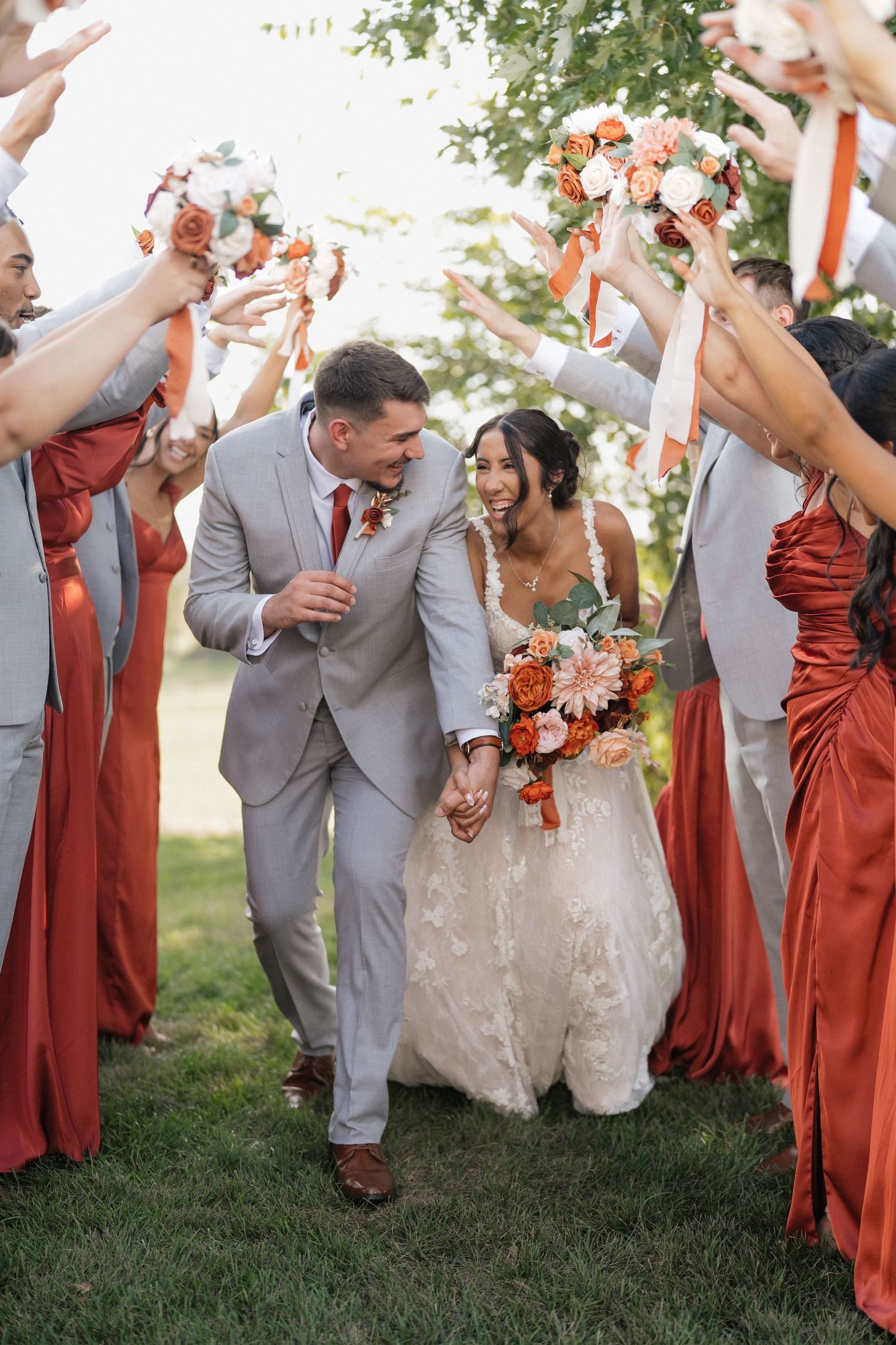 Wedding party celebrates outdoors with joyful poses in gray suits and rust-colored dresses.