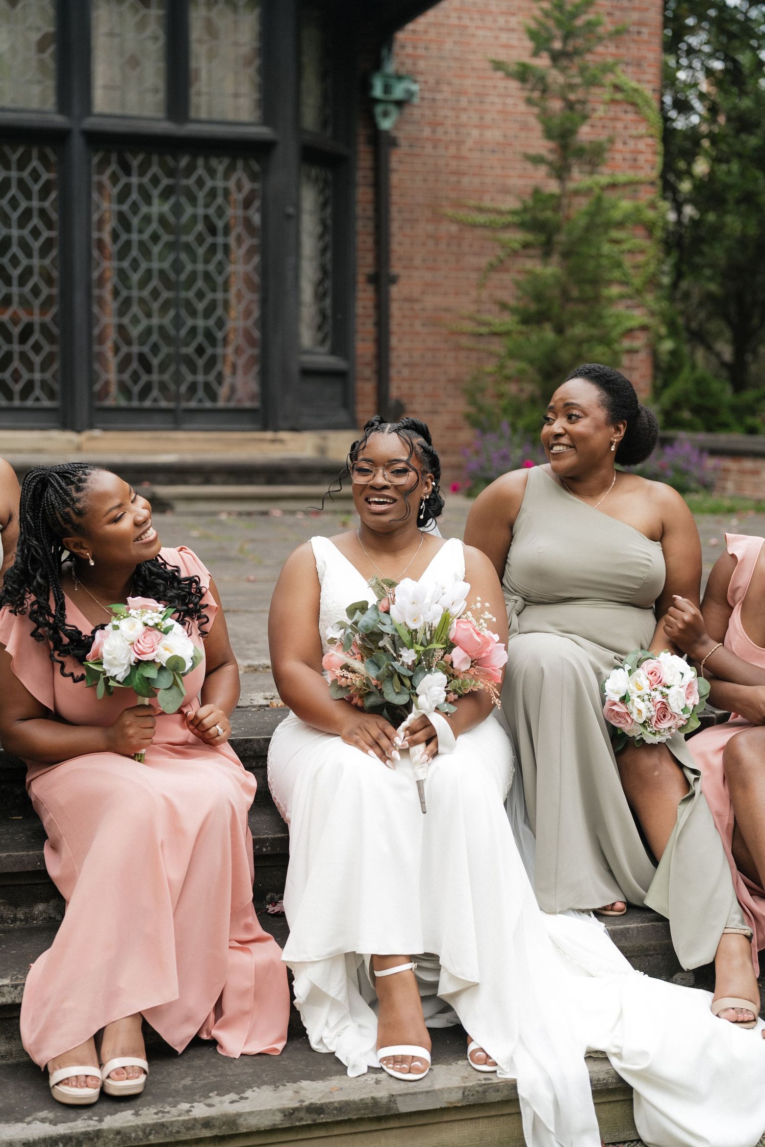 Wedding party seated outdoors sharing laughs with bouquets against brick building.