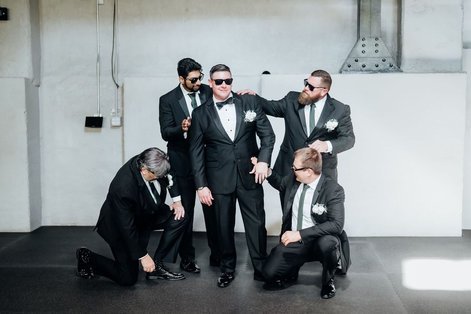 Group of formally dressed people in sunglasses posing playfully against a white wall.