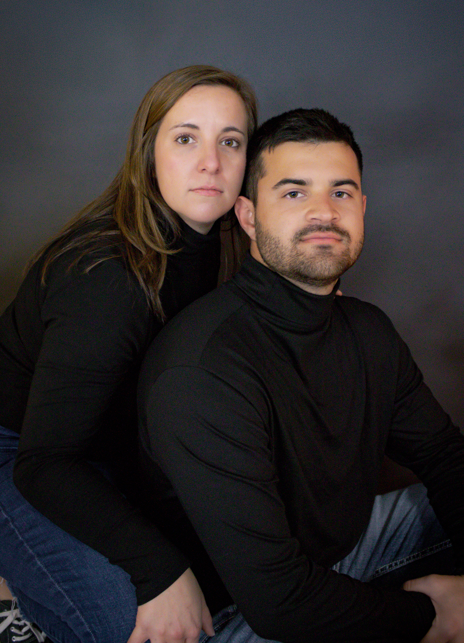 Couple in black clothing poses together against dark backdrop for intimate portrait session.
