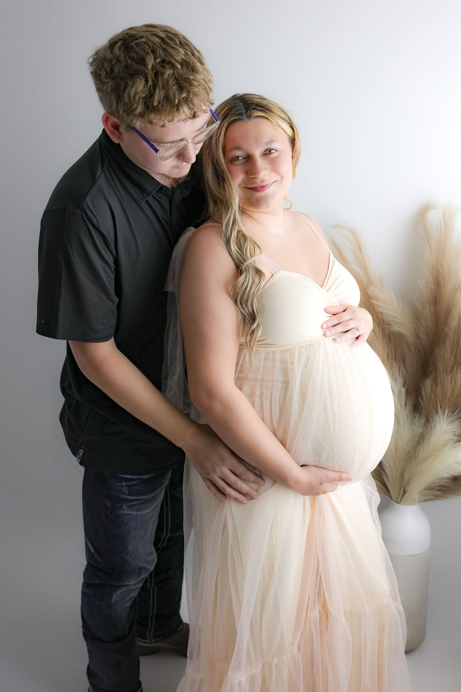 Maternity photo series showing tender moments between couple in light cream dress and black shirt against neutral background.