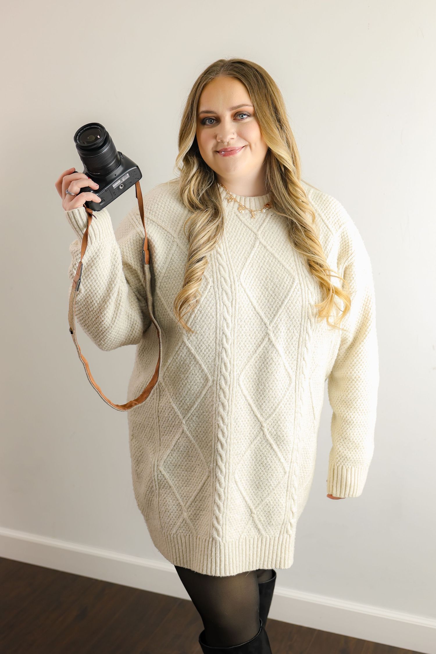 Woman in cream cable-knit sweater dress holding a camera with brown leather strap, smiling against white wall.