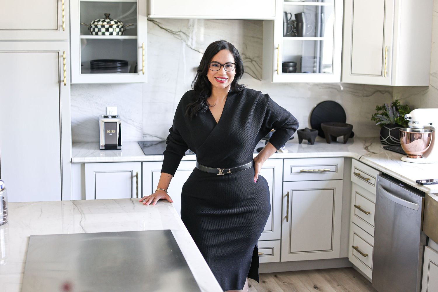 Woman in black dress posing confidently in a modern white kitchen with marble countertops and stainless appliances.
