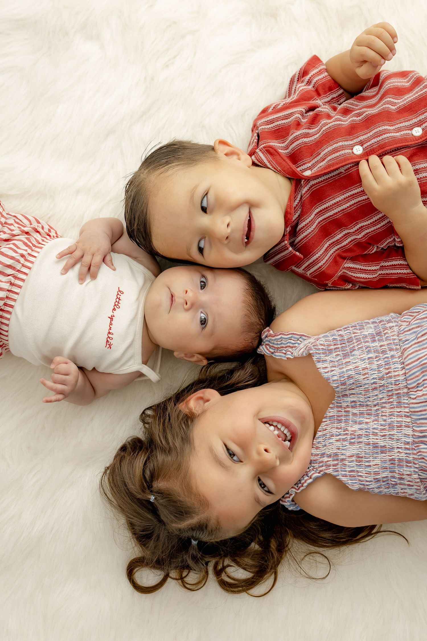 Three young siblings lying on a white fluffy rug, smiling together in red and blue outfits.