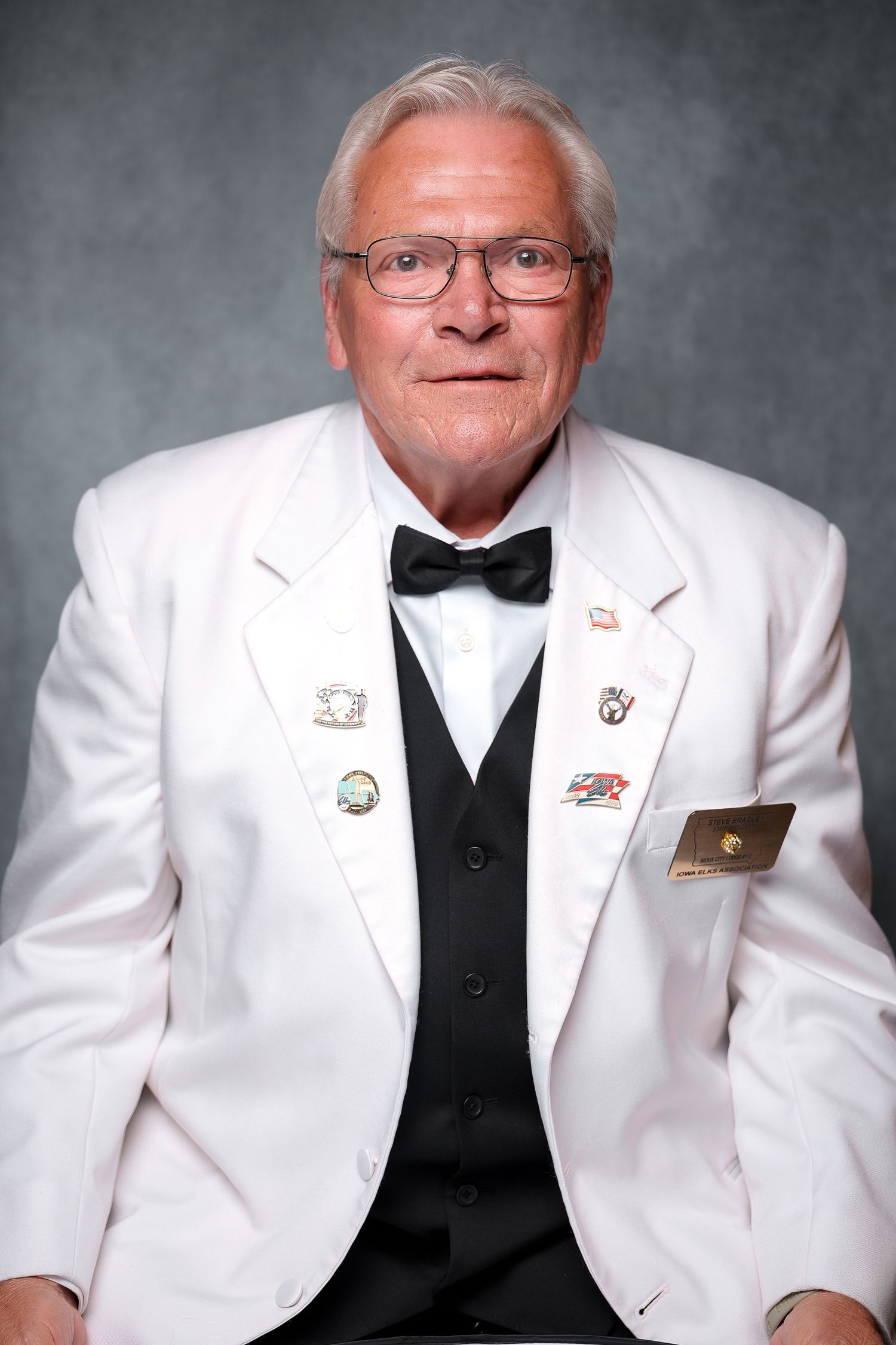 Elderly man in white blazer with pins, black bow tie and vest, wearing glasses, formal portrait photo.
