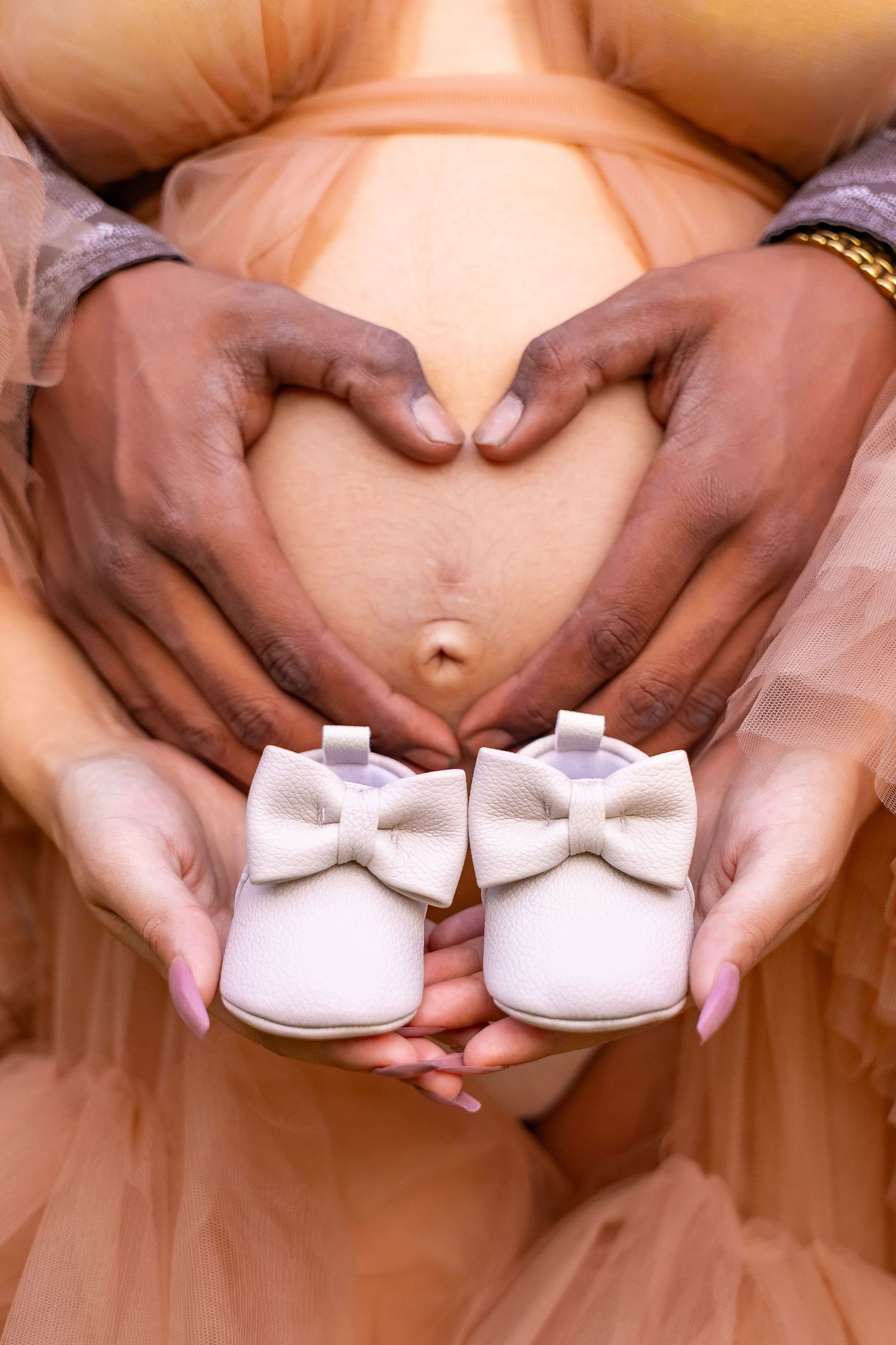 Hands form a heart shape over a pregnant belly while holding tiny white baby shoes with purple bows.