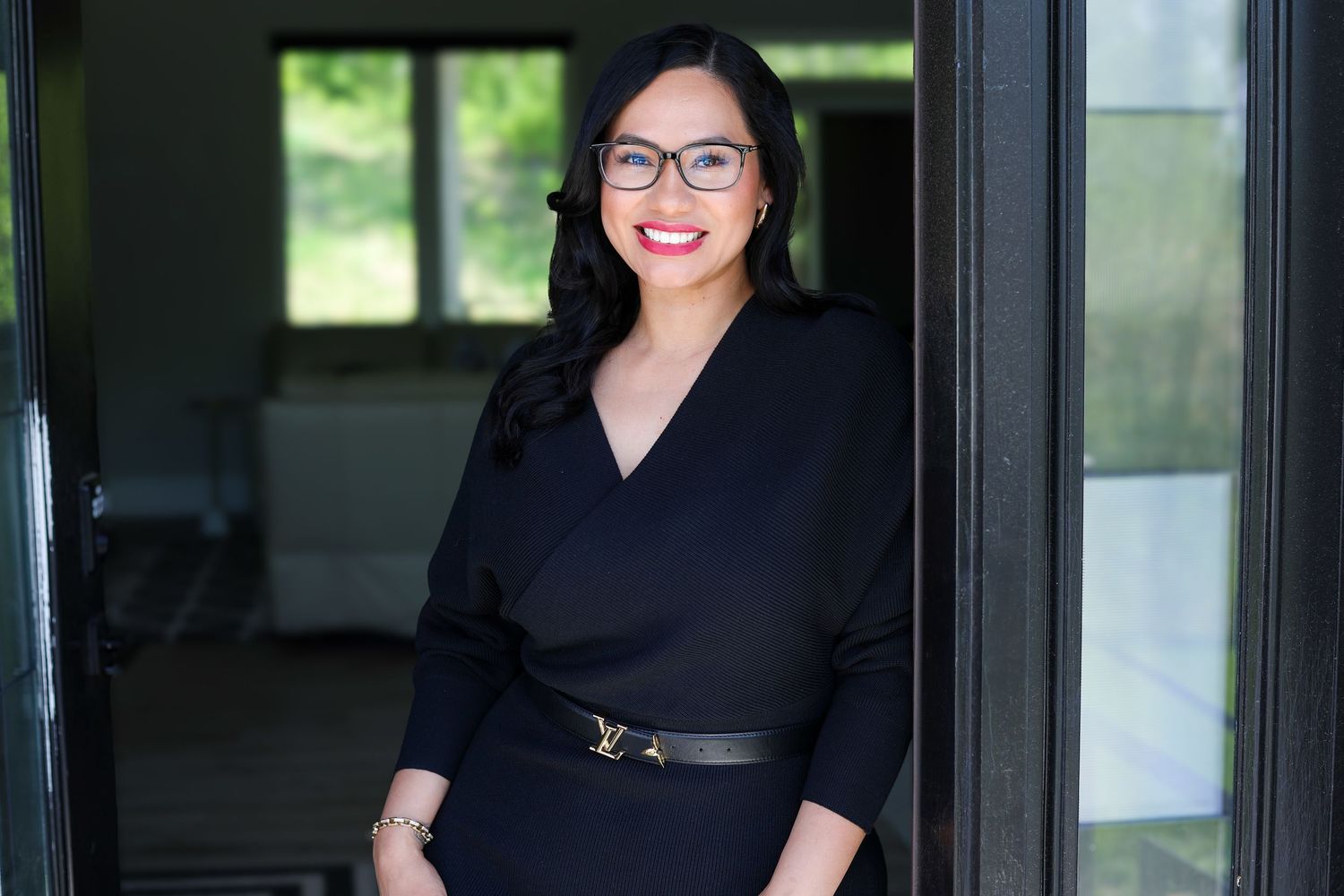 Professional woman in black dress and glasses smiling confidently in a modern doorway setting.