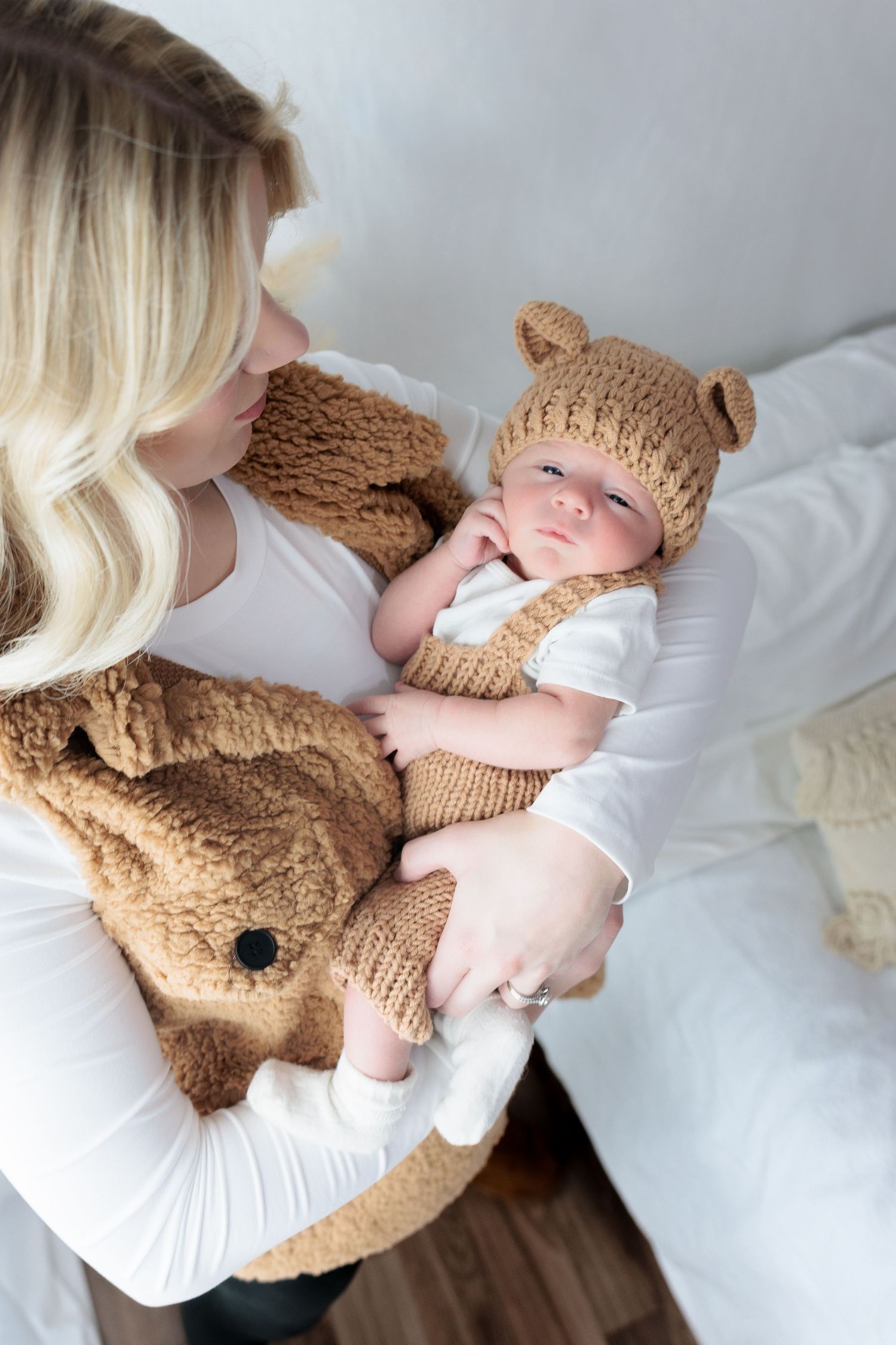 Baby wearing brown knit hat lies on white bedding while cuddling with plush teddy bear.