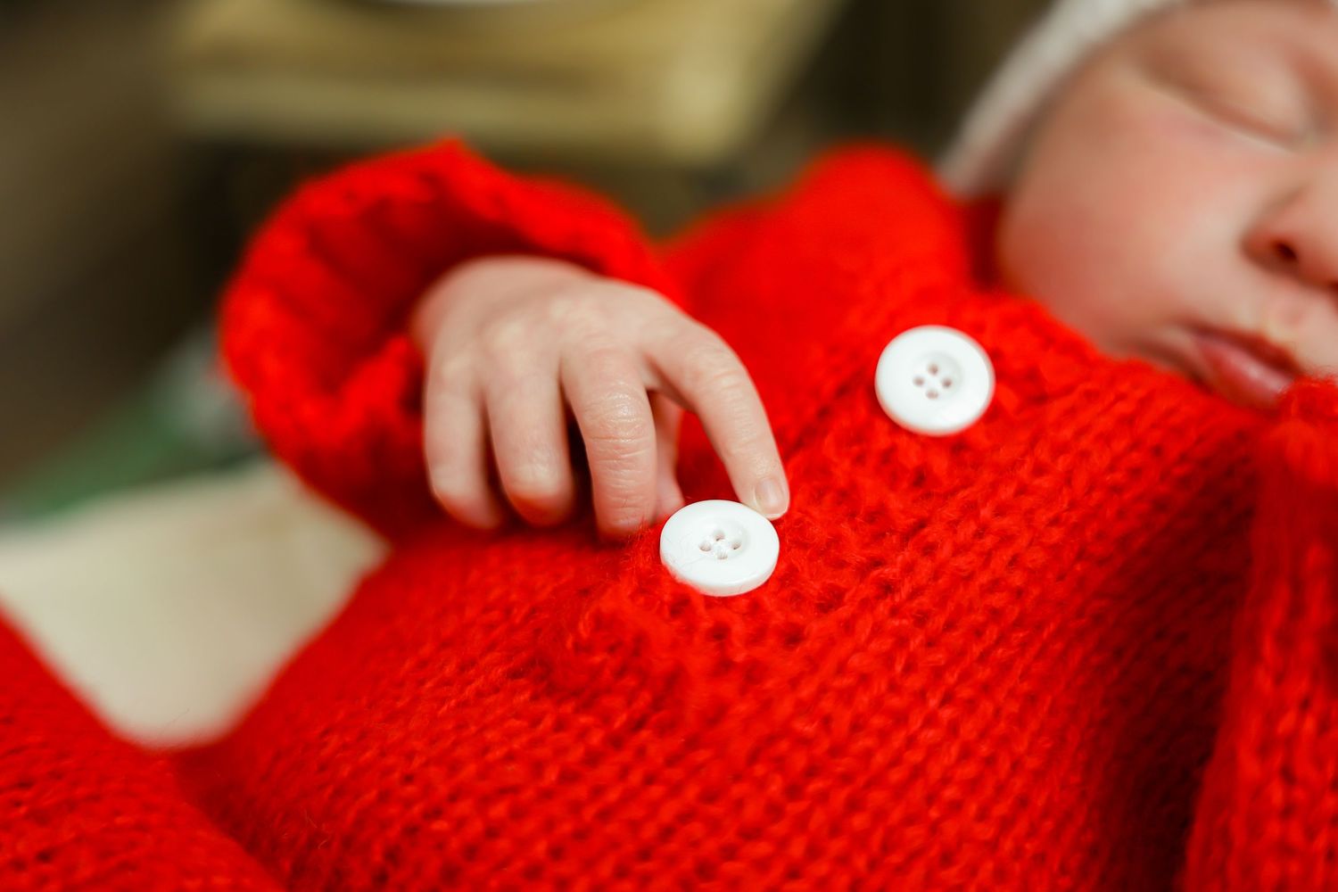 Close-up details of red knitted baby clothing with white buttons against plaid fabric background.