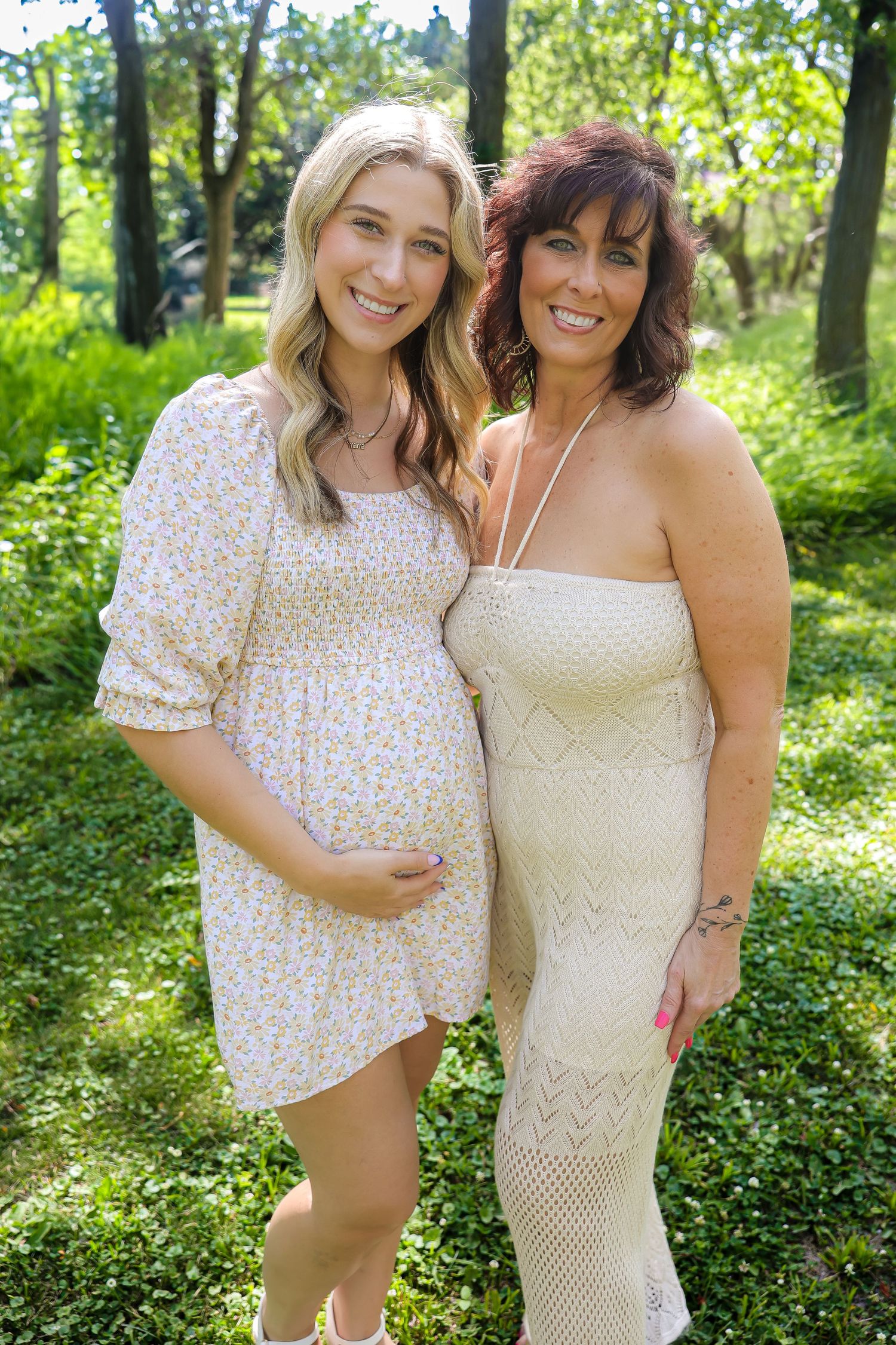 Two people wearing light colored dresses stand together in a wooded garden setting.