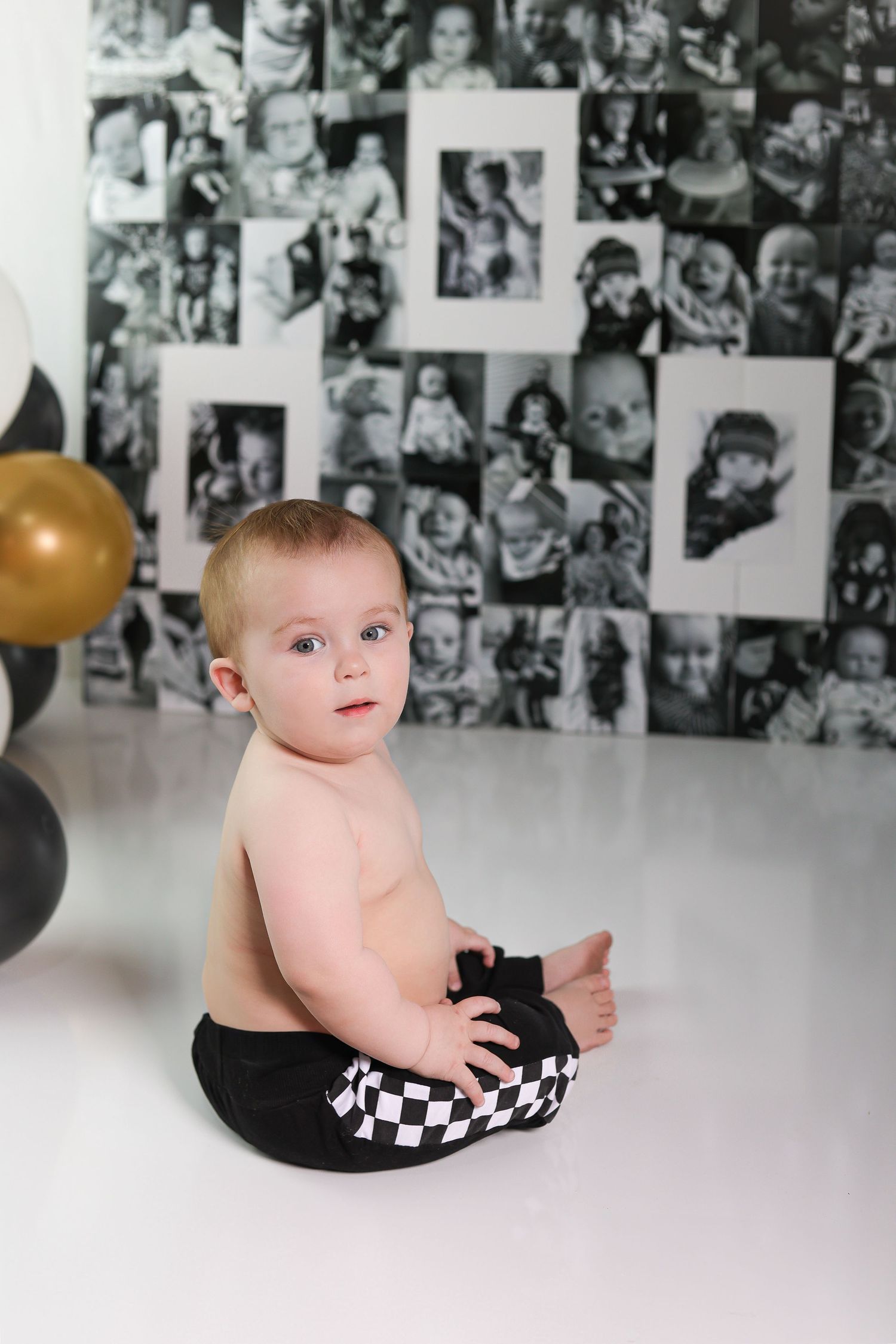 A baby in checkered pants poses in front of a photo collage wall with balloons.