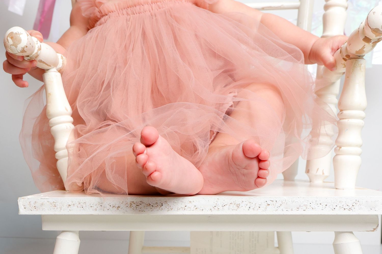 Tiny feet in pink tulle skirt peek out while resting on white wooden chair.