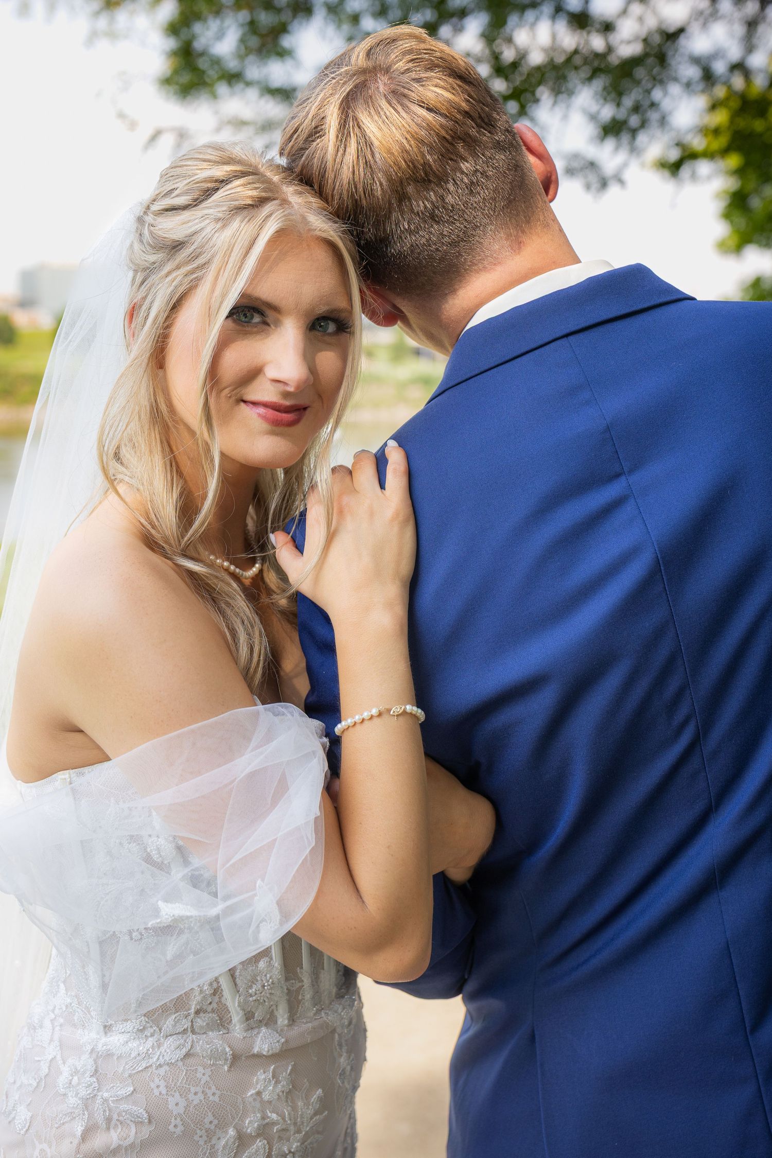 Romantic wedding couple embraces outdoors, bride in white lace dress and groom in navy suit share intimate moment.