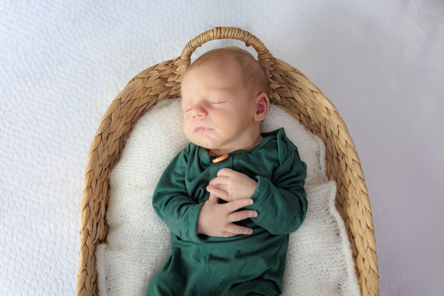 A sleeping baby in a woven basket wearing a dark green outfit lies on a white textured blanket.