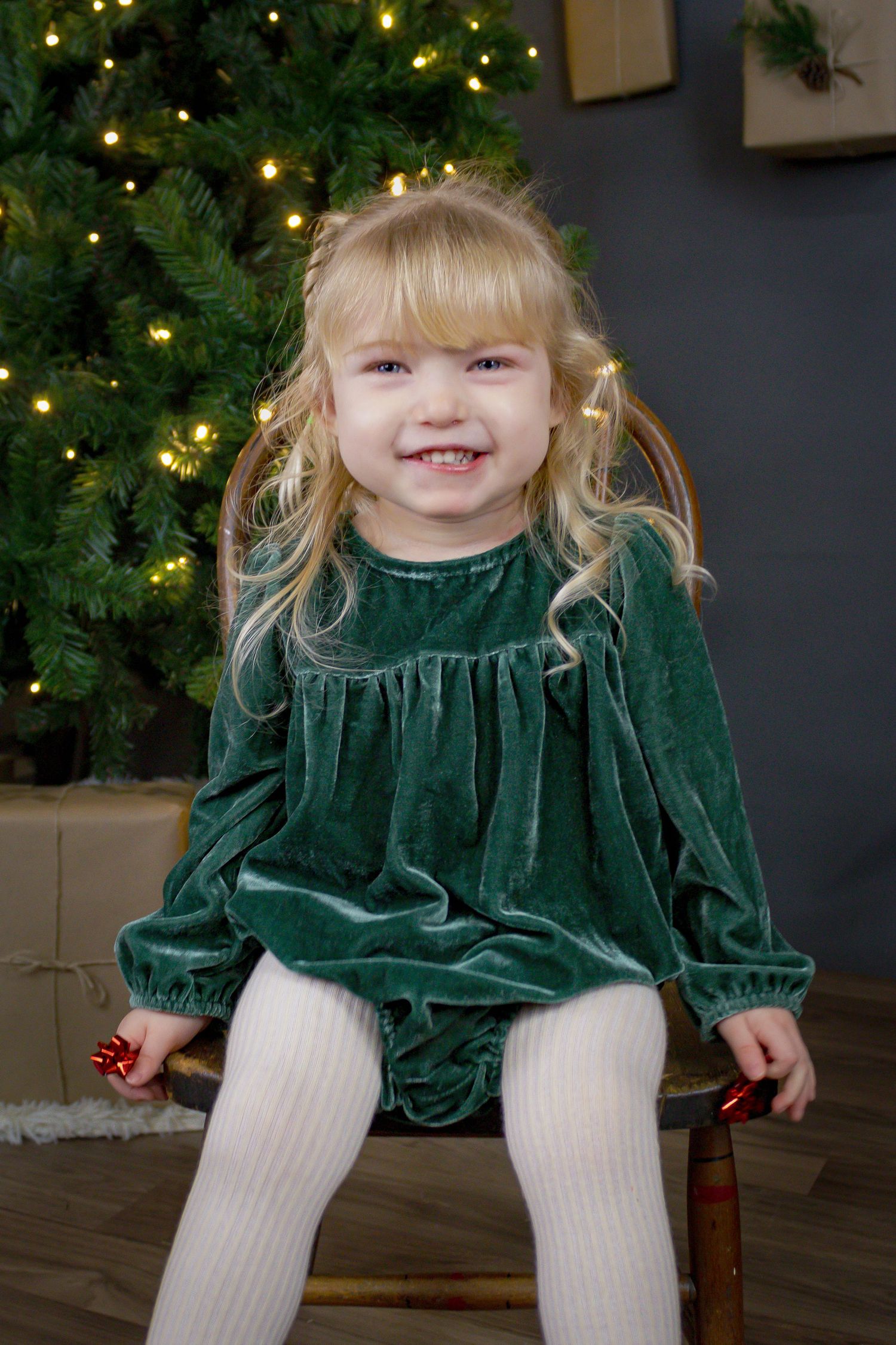 Child in emerald velvet dress sits in front of decorated Christmas tree with twinkling lights.