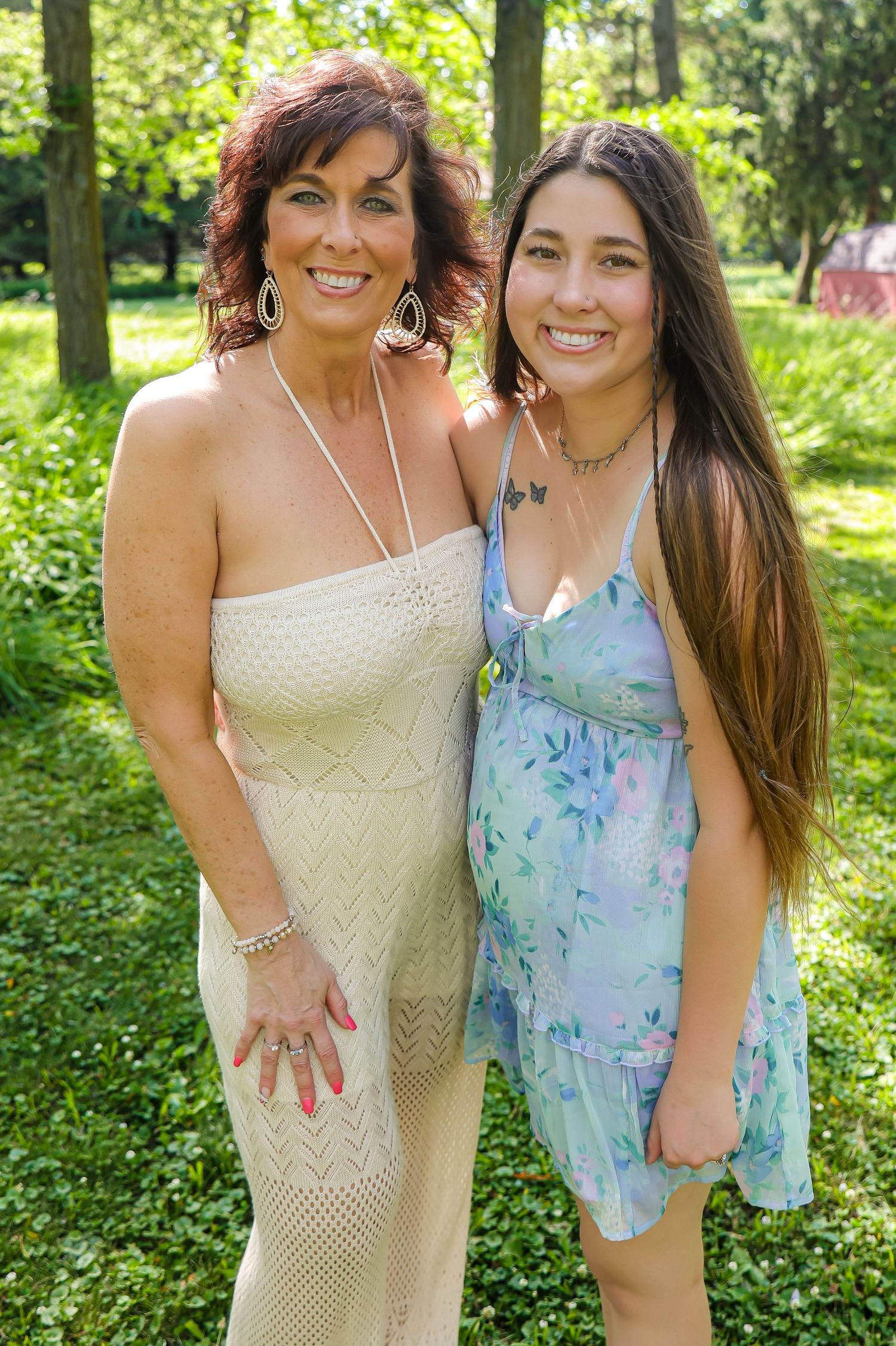 Two people in summer dresses smile together in front of green foliage.