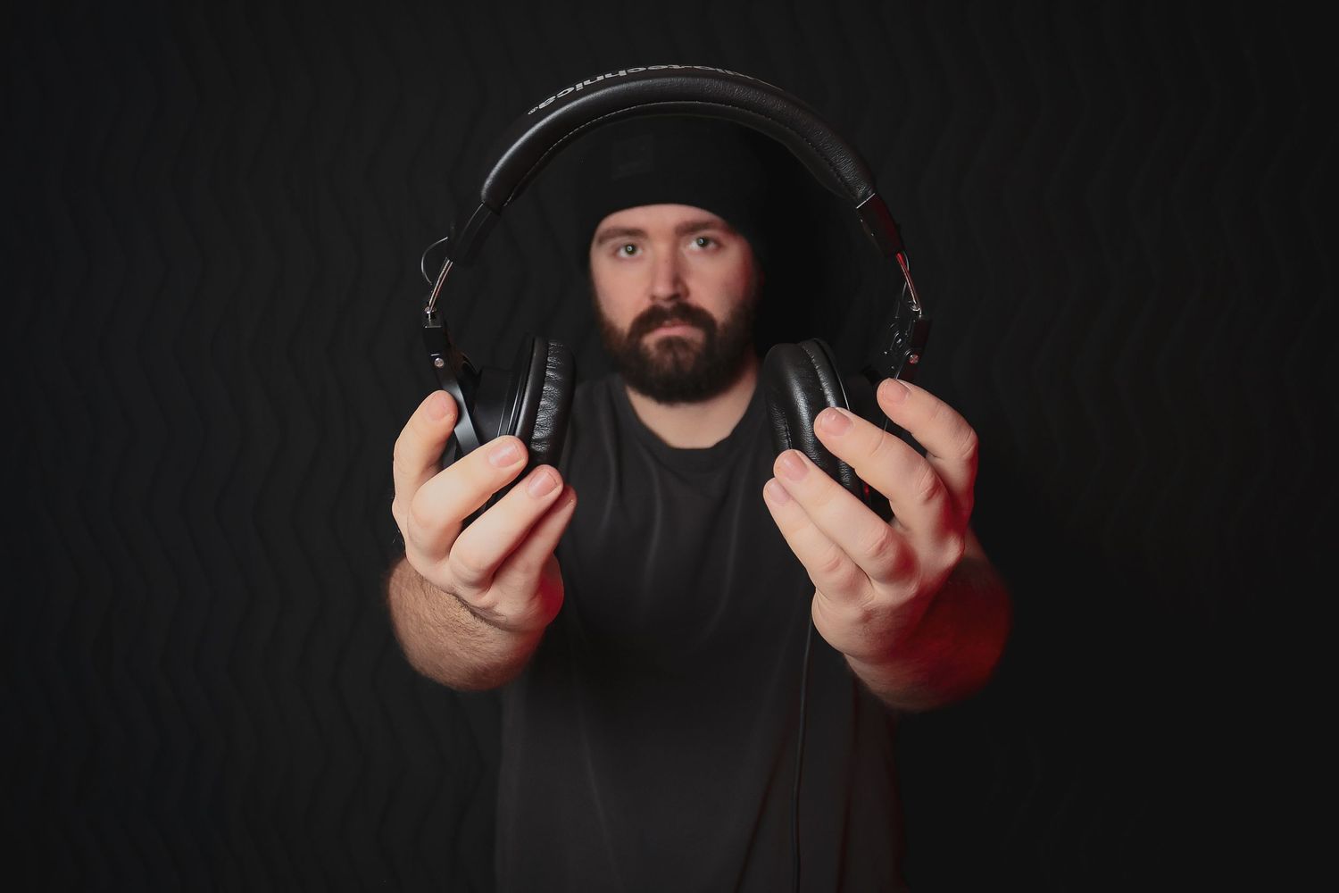 Person holding professional headphones against dark background while wearing black clothing.