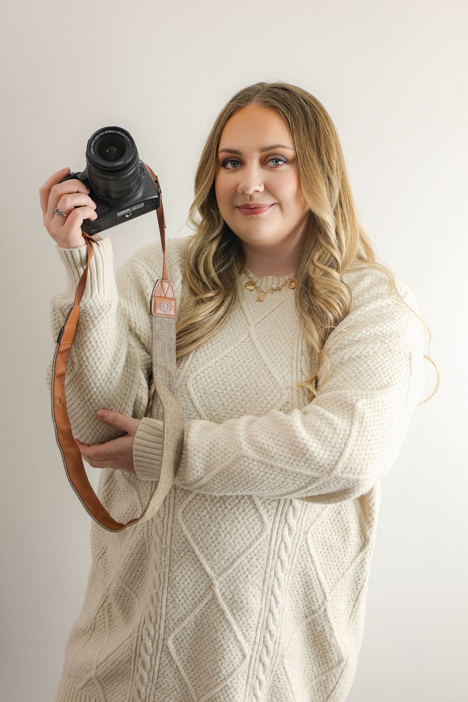 Smiling woman in cream knit sweater holding camera with leather strap, posing against light background.