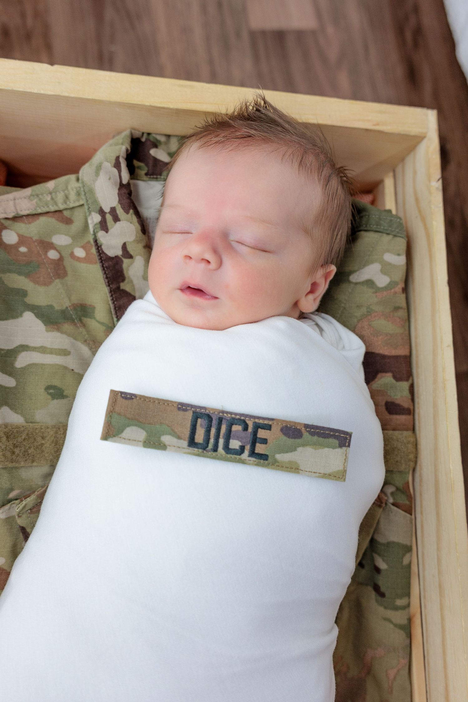 A sleeping newborn baby wearing military camouflage and white clothing in a wooden crate.