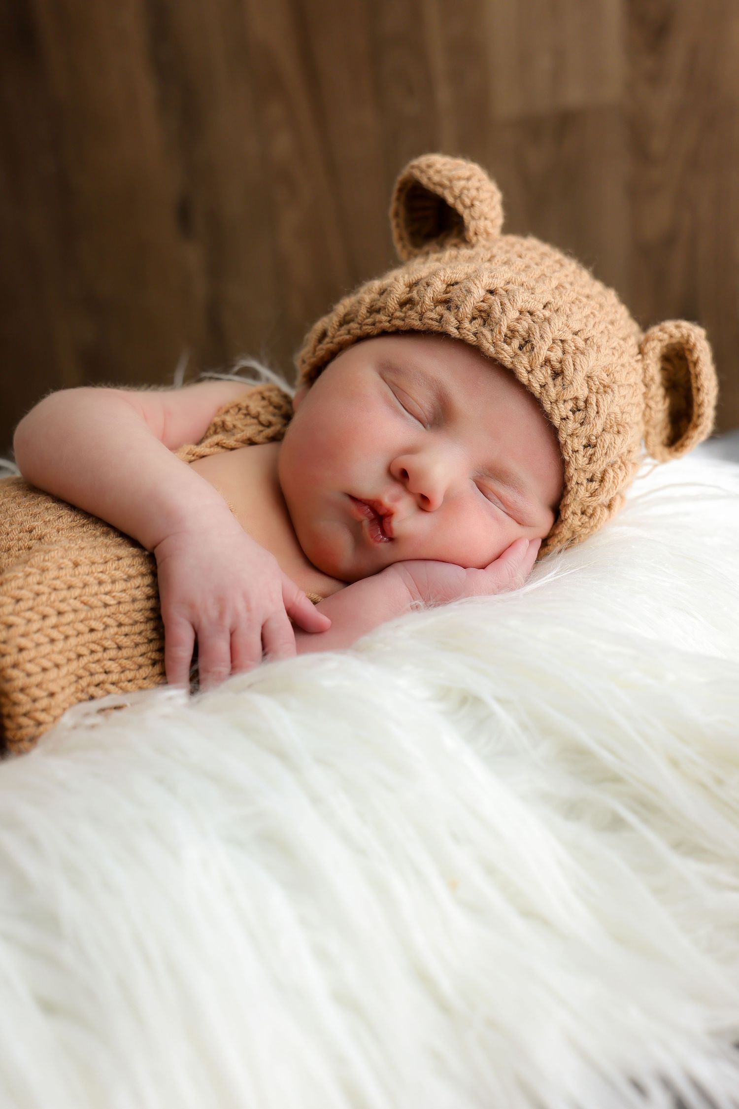 Sleeping baby wearing a cozy brown knit bear hat nestled in white fluffy blankets.