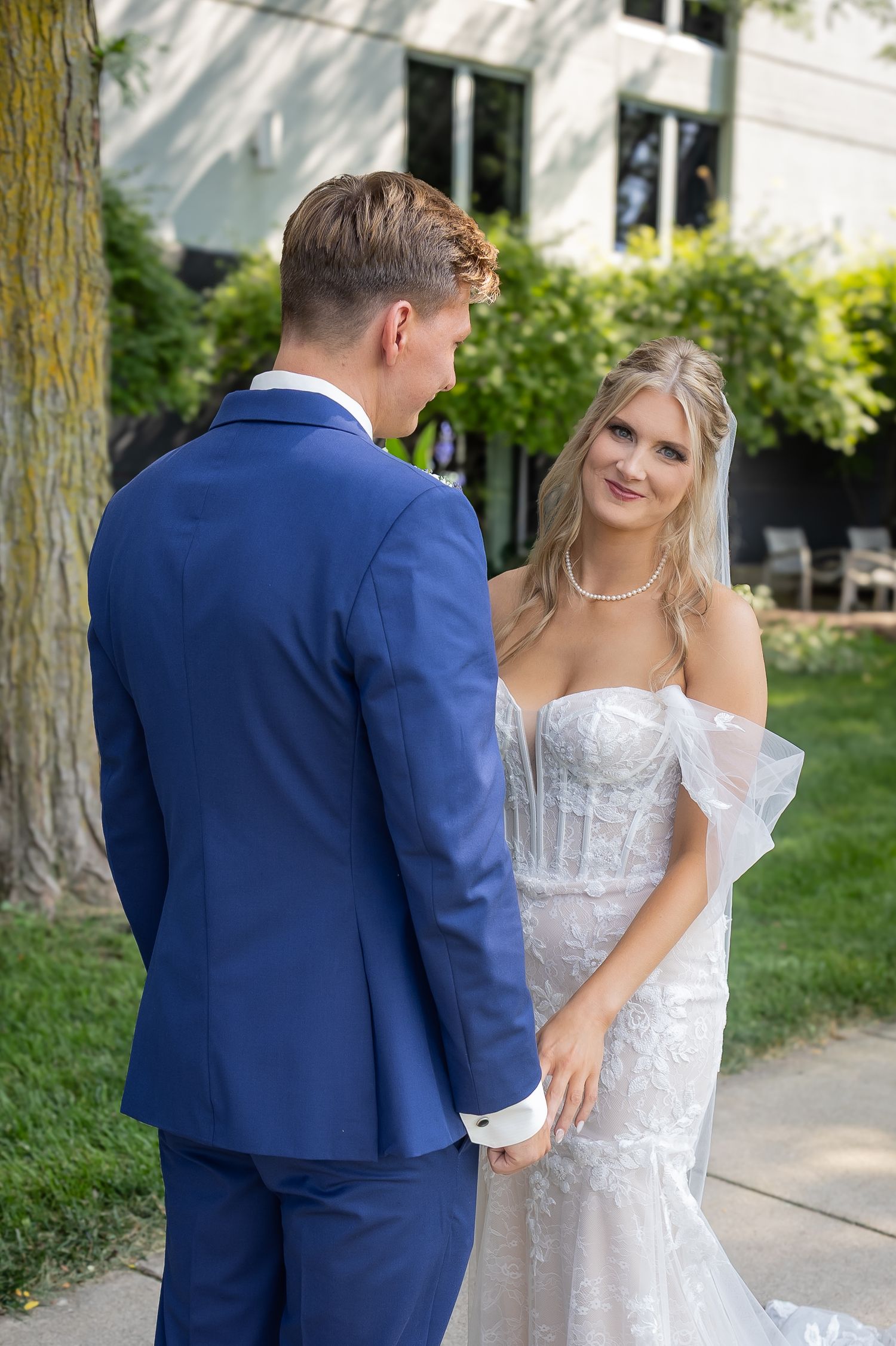 A bride in a white lace gown and groom in navy suit share a moment together on their wedding day in a garden setting.