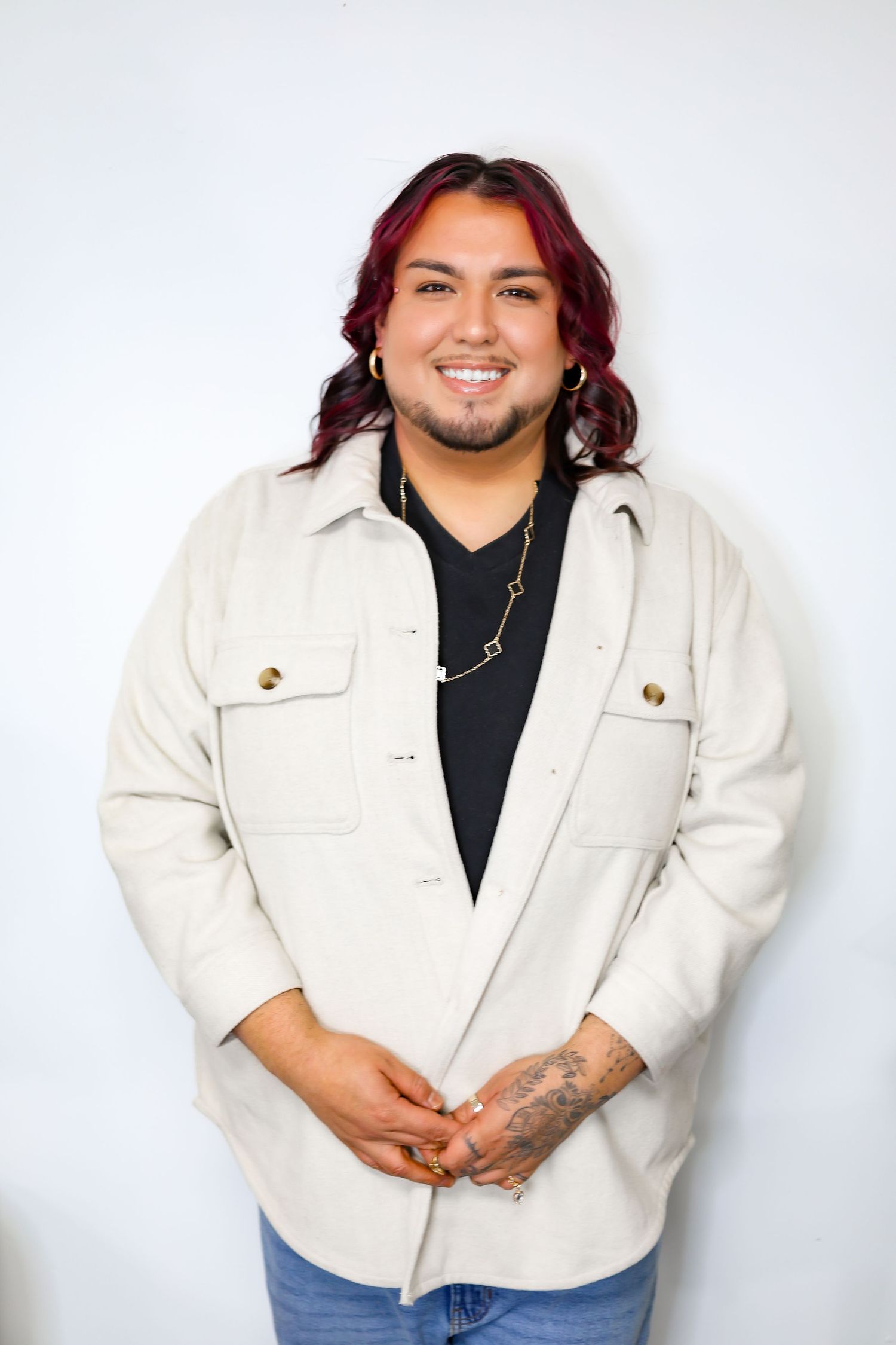 Professional hair stylist wearing white denim jacket posing in modern studio setting.