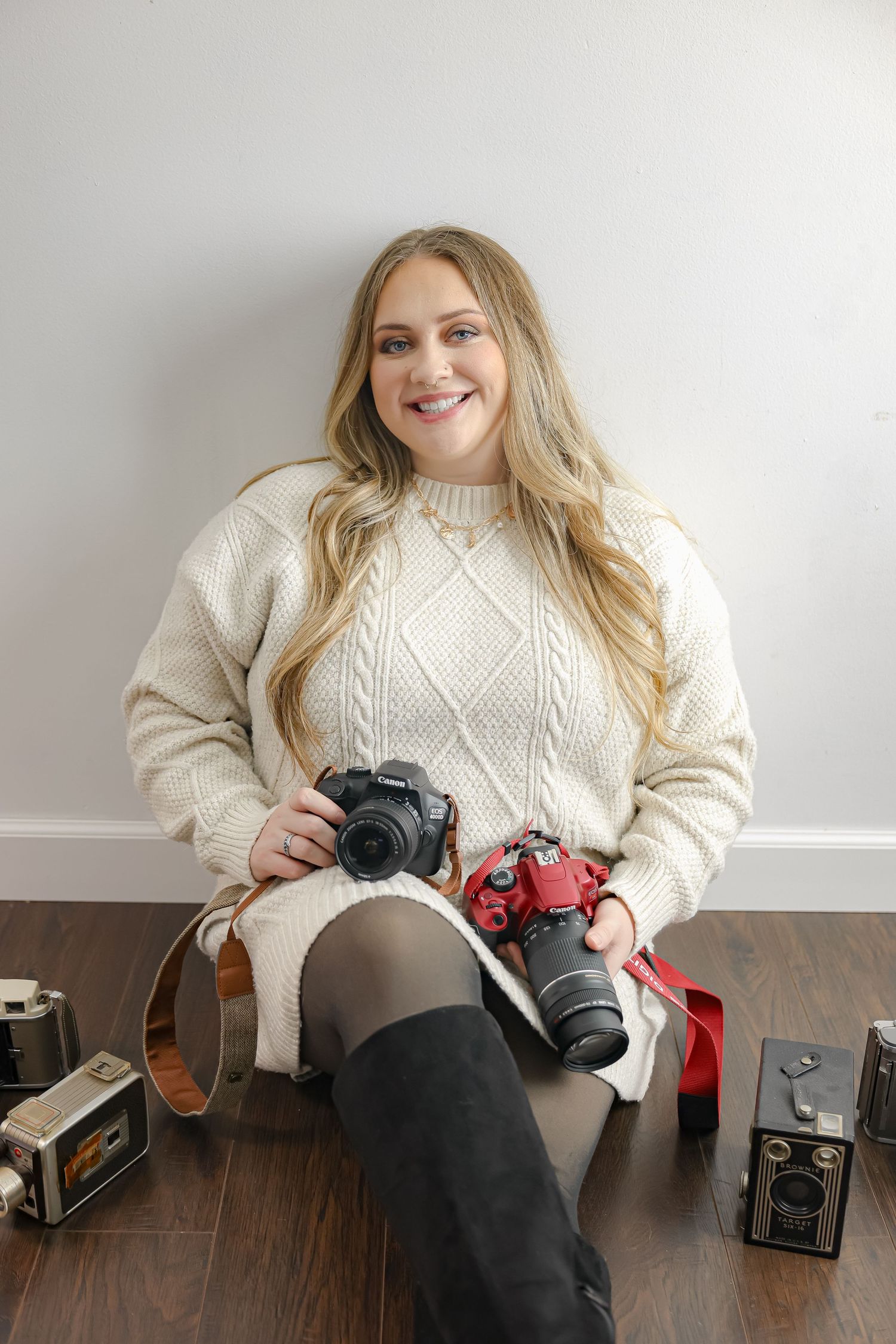 Smiling blonde woman in cream cable-knit sweater sitting on floor holding two DSLR cameras surrounded by vintage cameras.