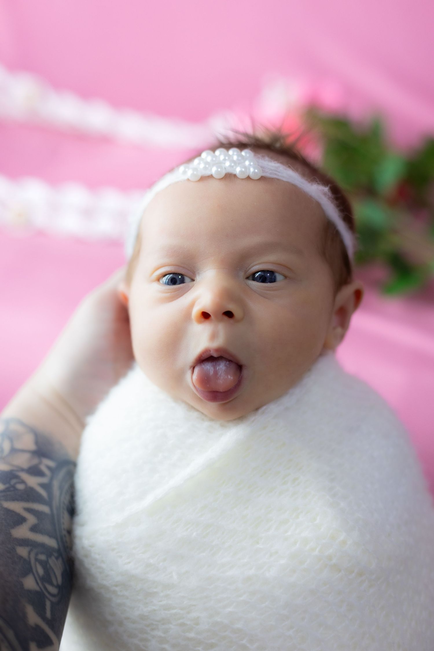 Playful newborn portrait with white wrap and headband against pink background.