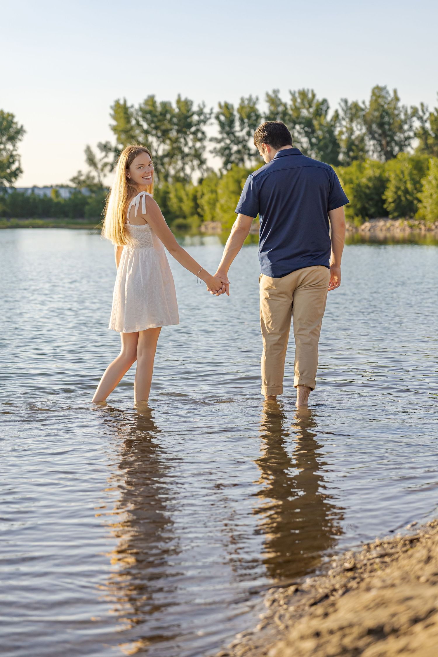 Two people wading in shallow lake water at sunset with trees in the background.
