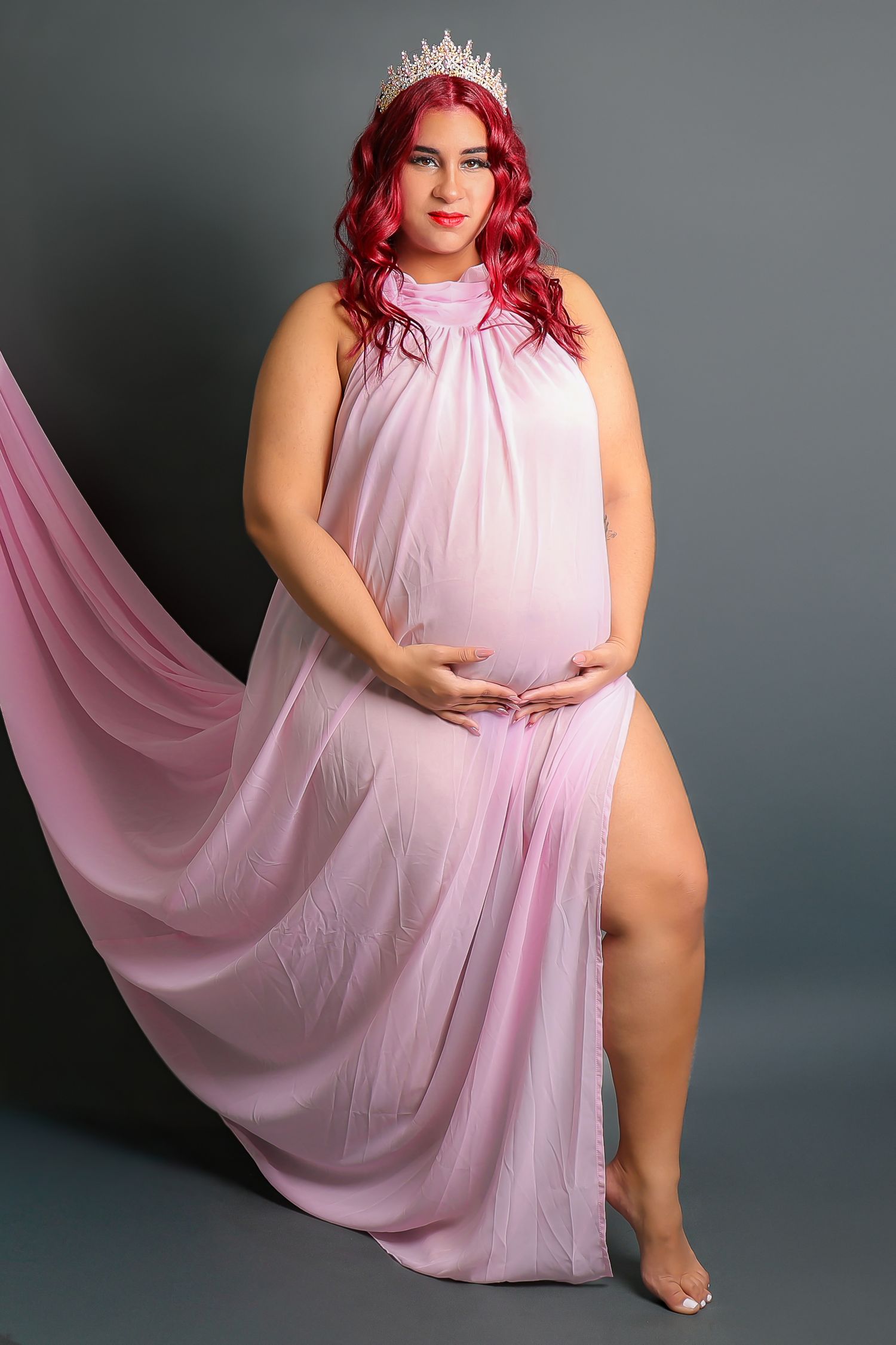Woman in flowing pink dress poses gracefully on dark backdrop wearing crown and showing elegant movements.