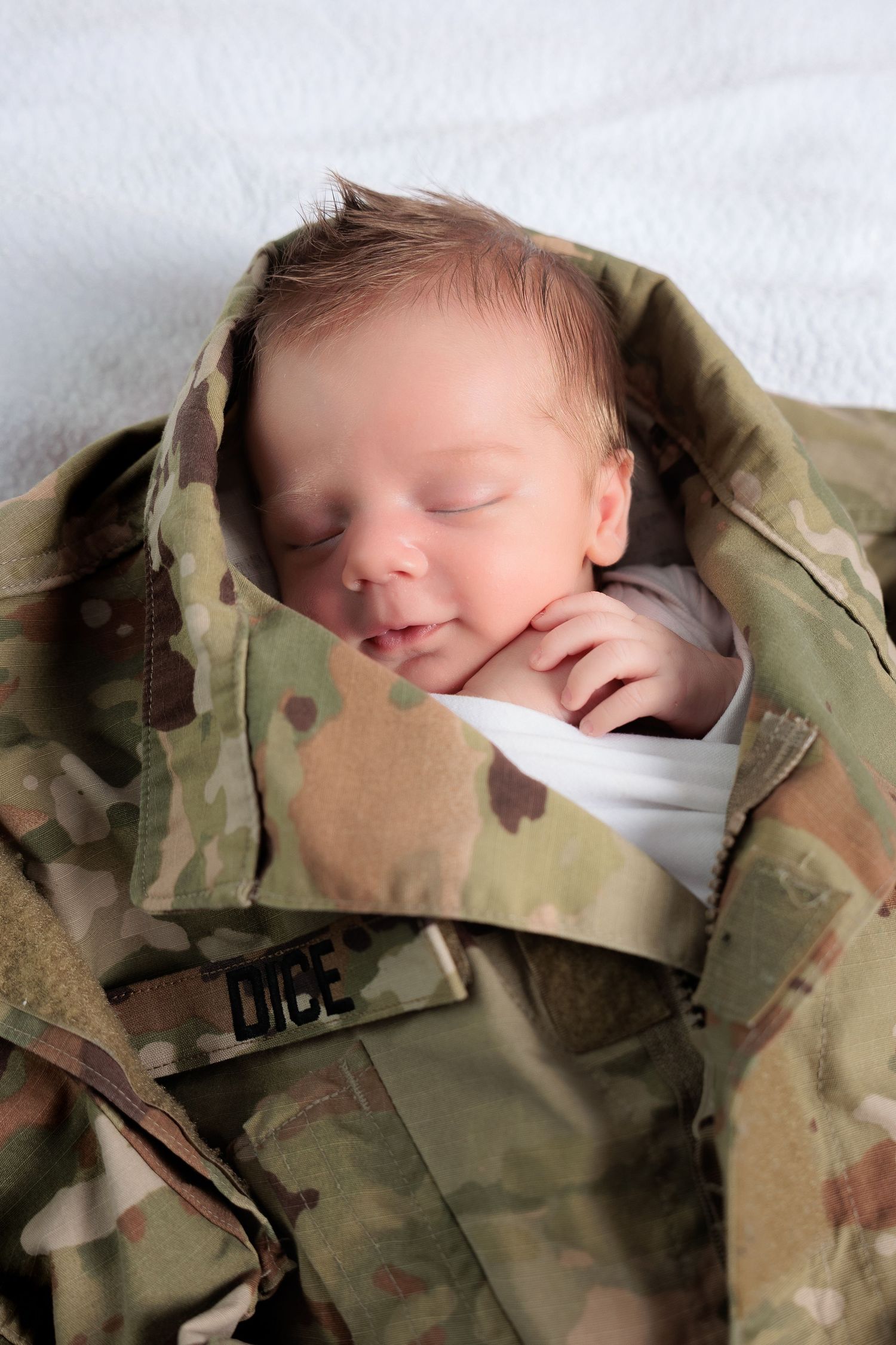 A sleeping baby wrapped in a camouflage military uniform jacket lies peacefully on white bedding.