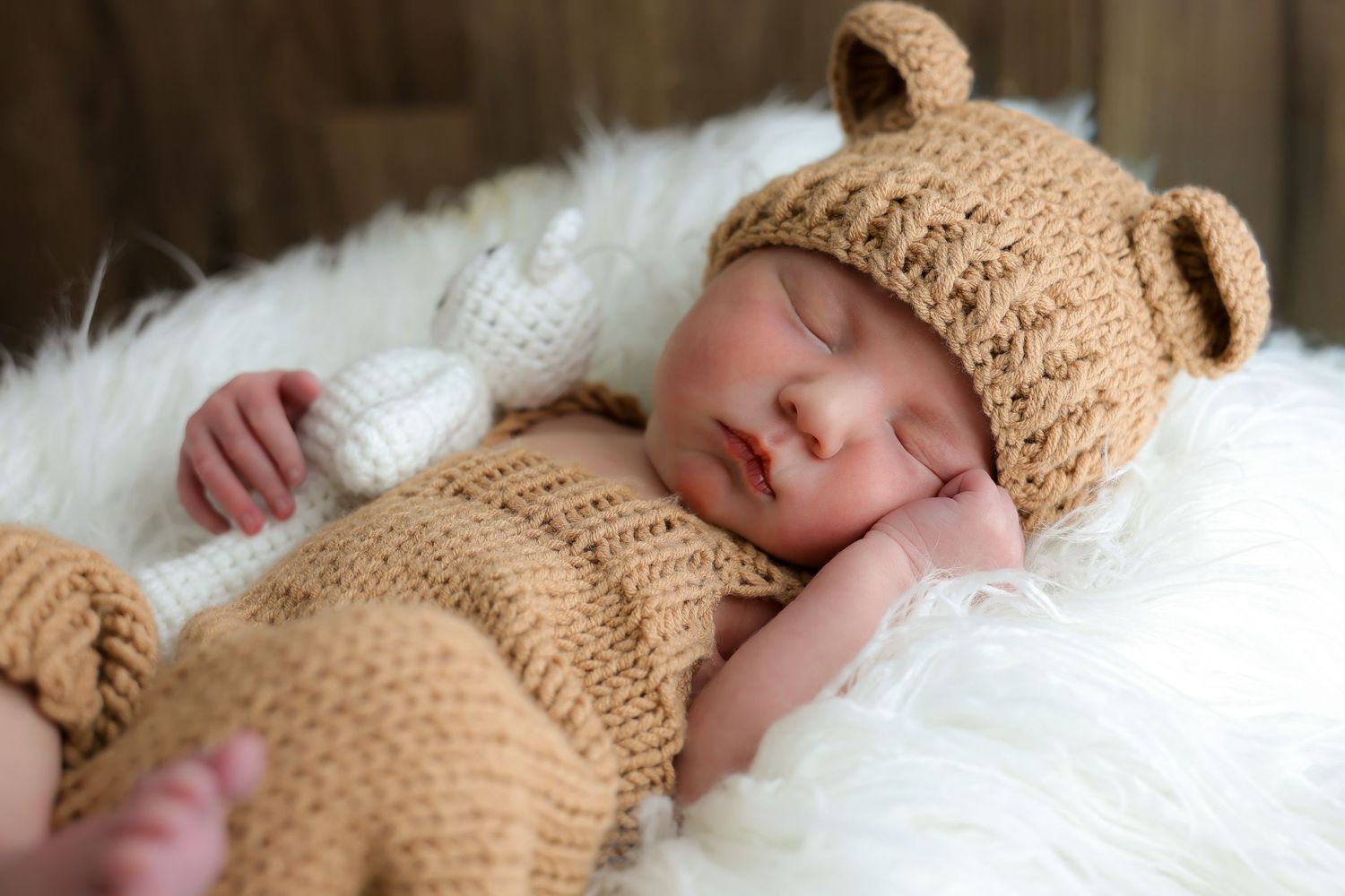 Sleeping newborn baby wearing knitted tan bonnet rests peacefully on soft white blanket.