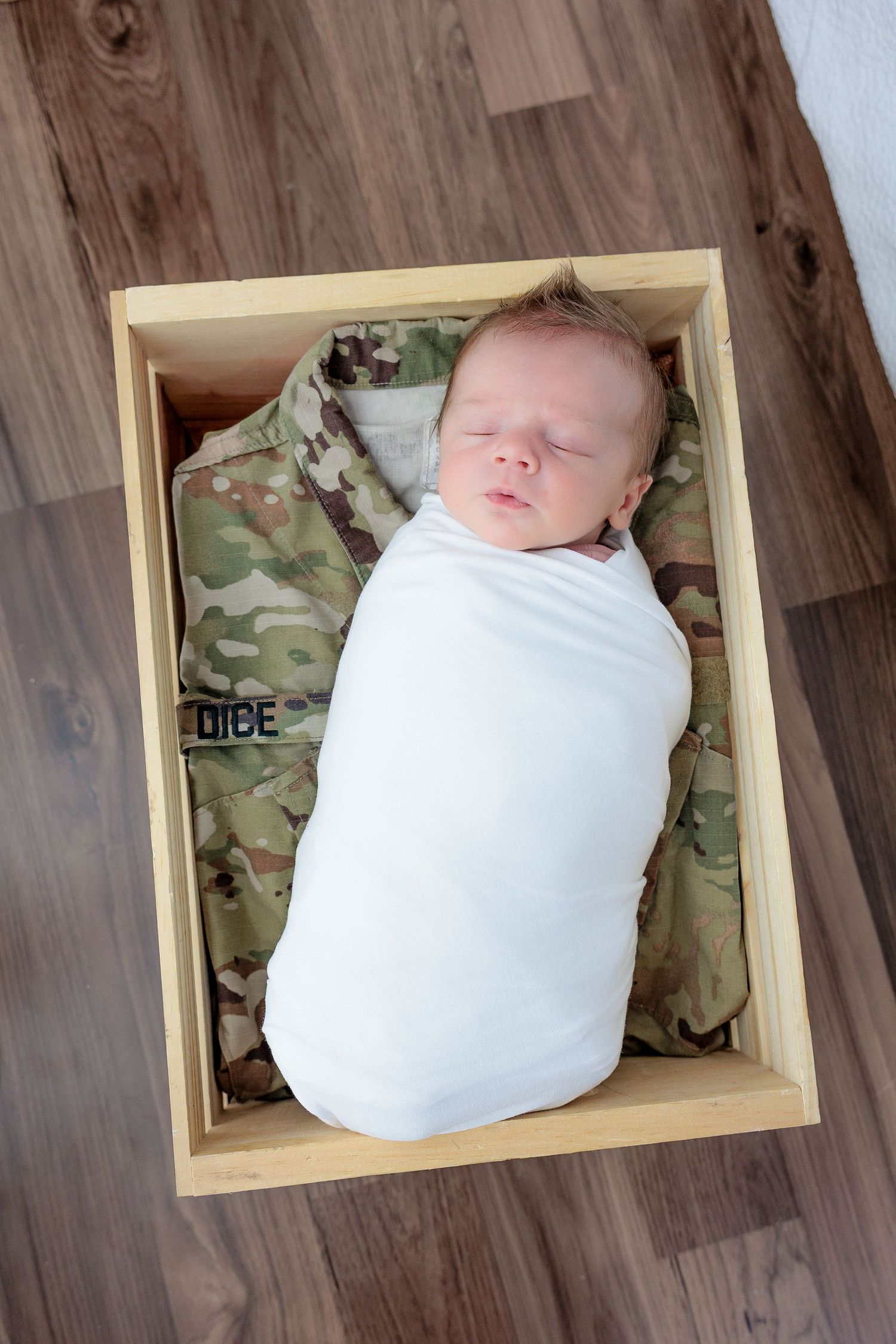 Newborn baby swaddled in white blanket rests peacefully in wooden box with camouflage fabric backdrop.