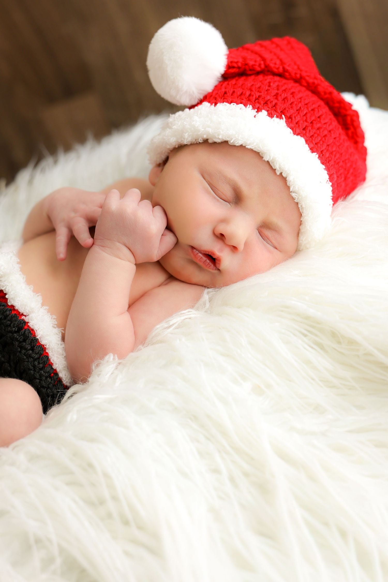 Peaceful sleeping baby wearing a red and white Santa hat rests on soft white fur.
