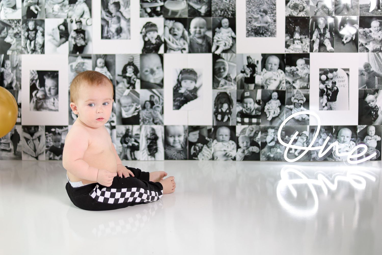 A baby in black and white checkered pants sits against a wall of vintage photographs.