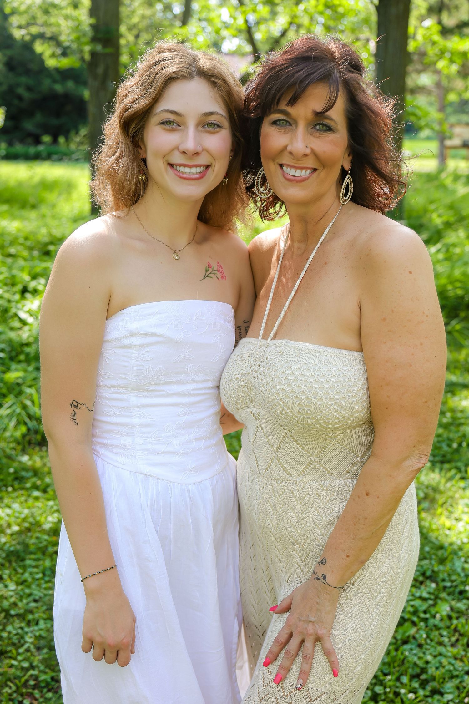 Two people in white and cream dresses pose together in a sunny garden.