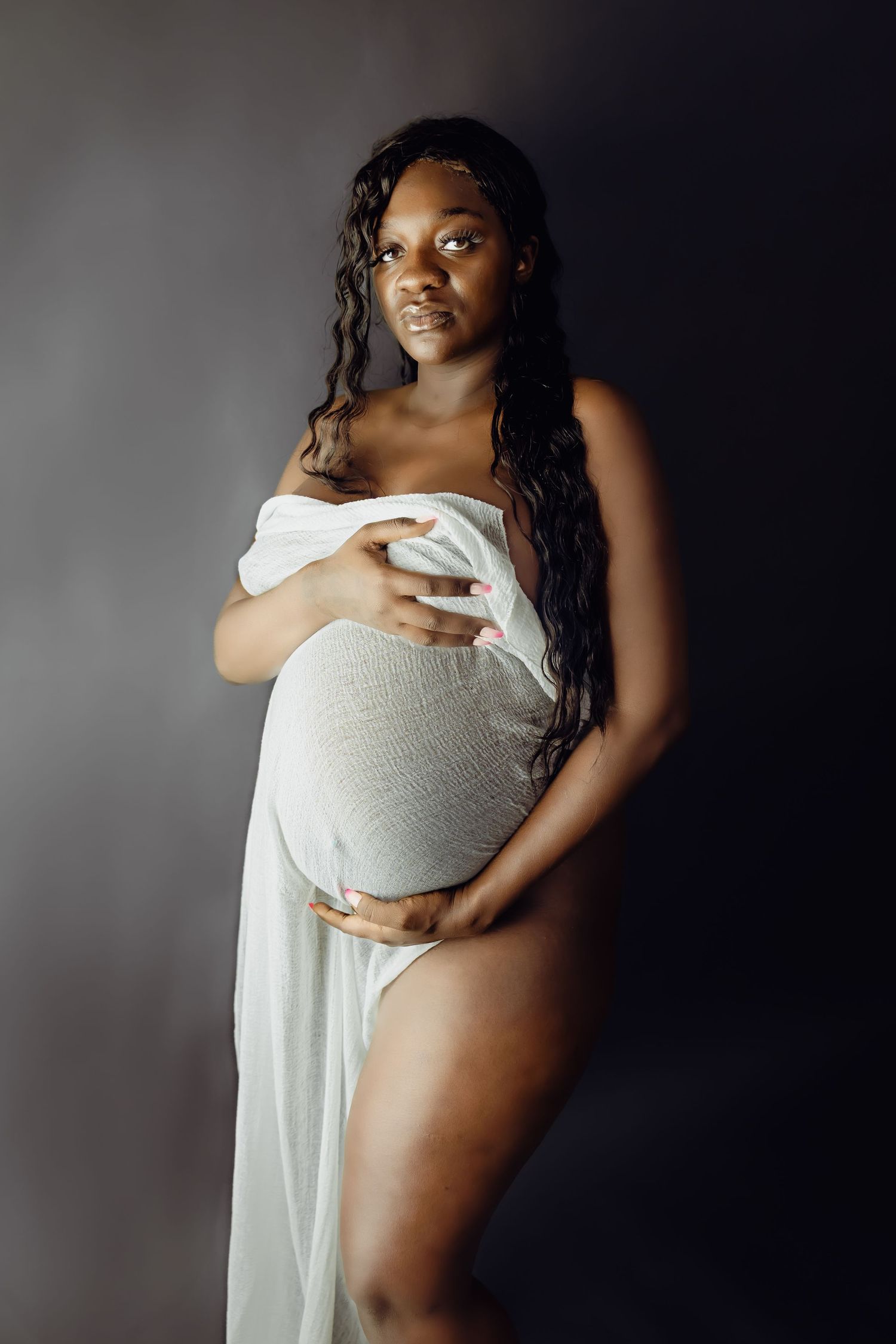 Artistic maternity photo of an expectant mother in white fabric against a dark backdrop.