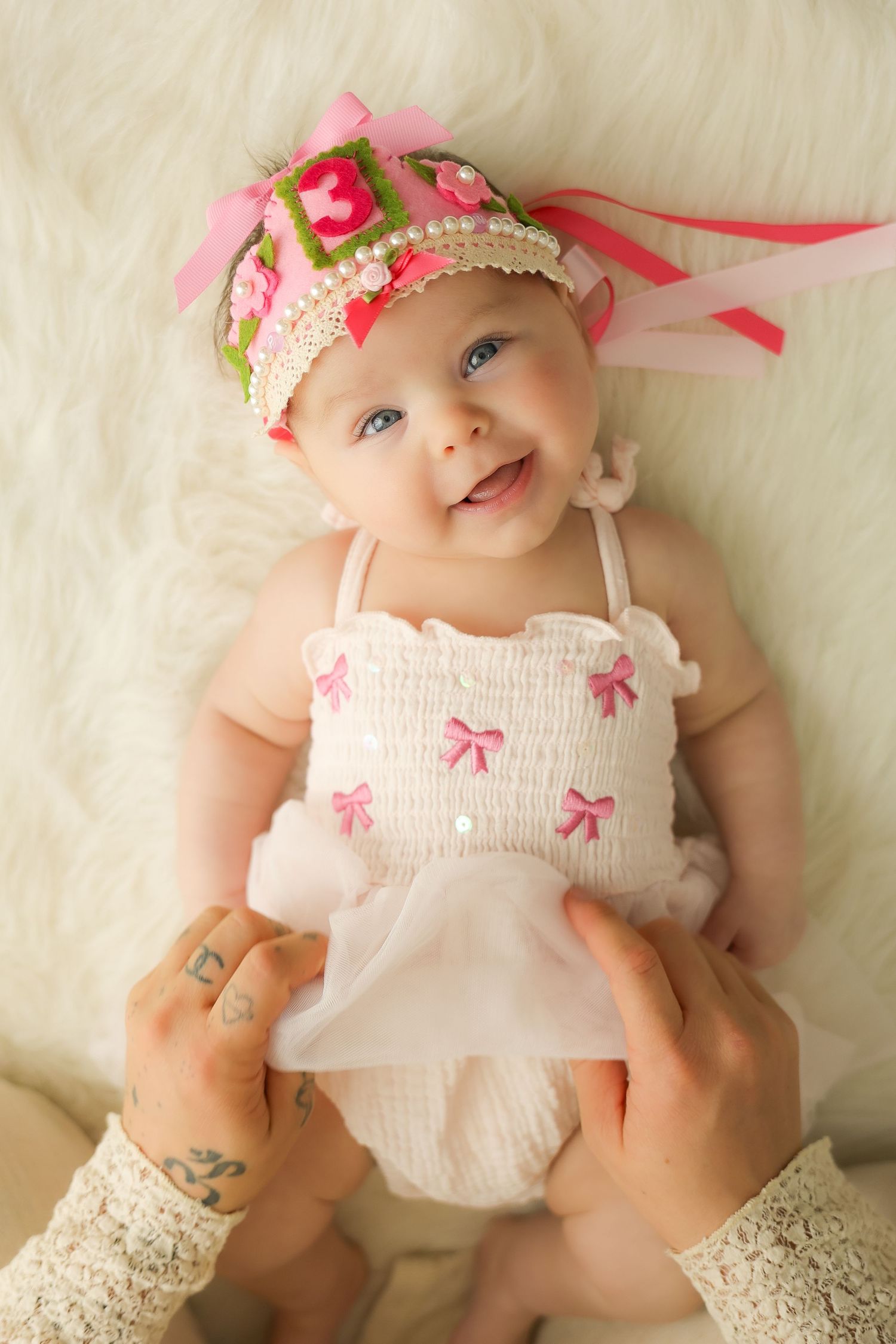 Smiling baby girl in white dress with pink bows and floral headband, held by tattooed hands on fluffy white background.