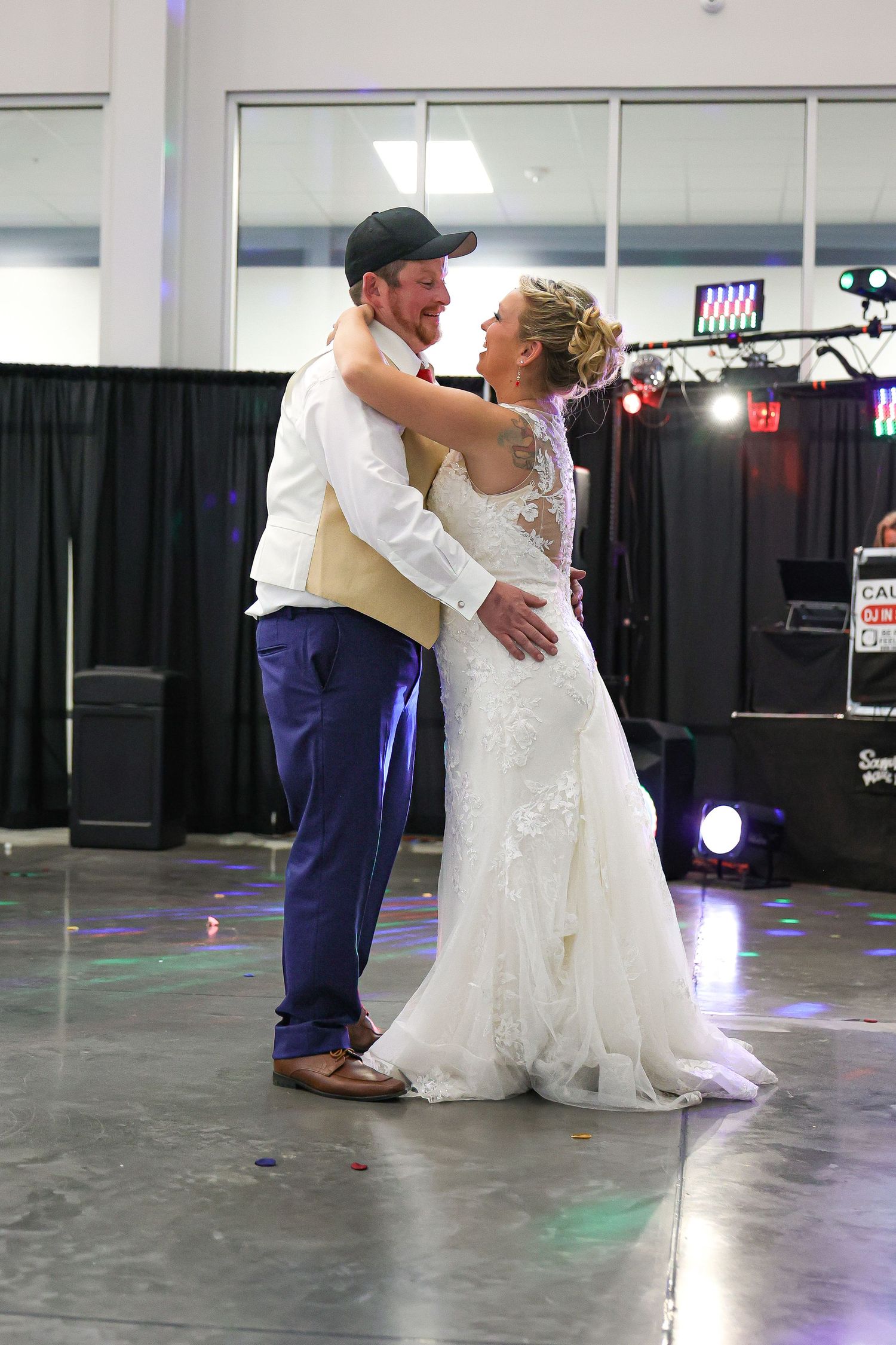 A couple shares their first dance at a wedding reception in a venue with colorful spotlights and a black curtain backdrop.