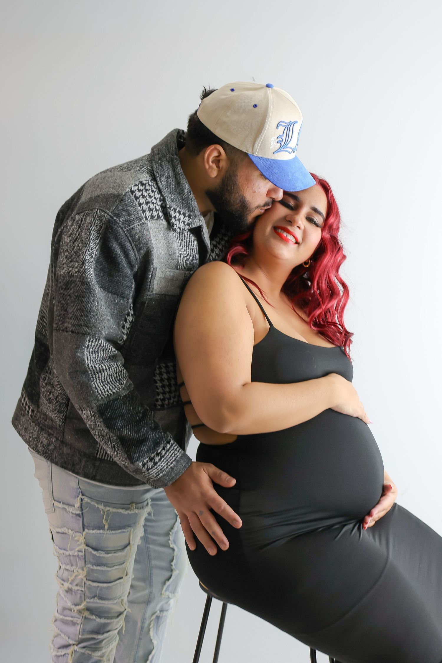 A person wearing a baseball cap and jeans leans over to embrace someone sitting in a chair.