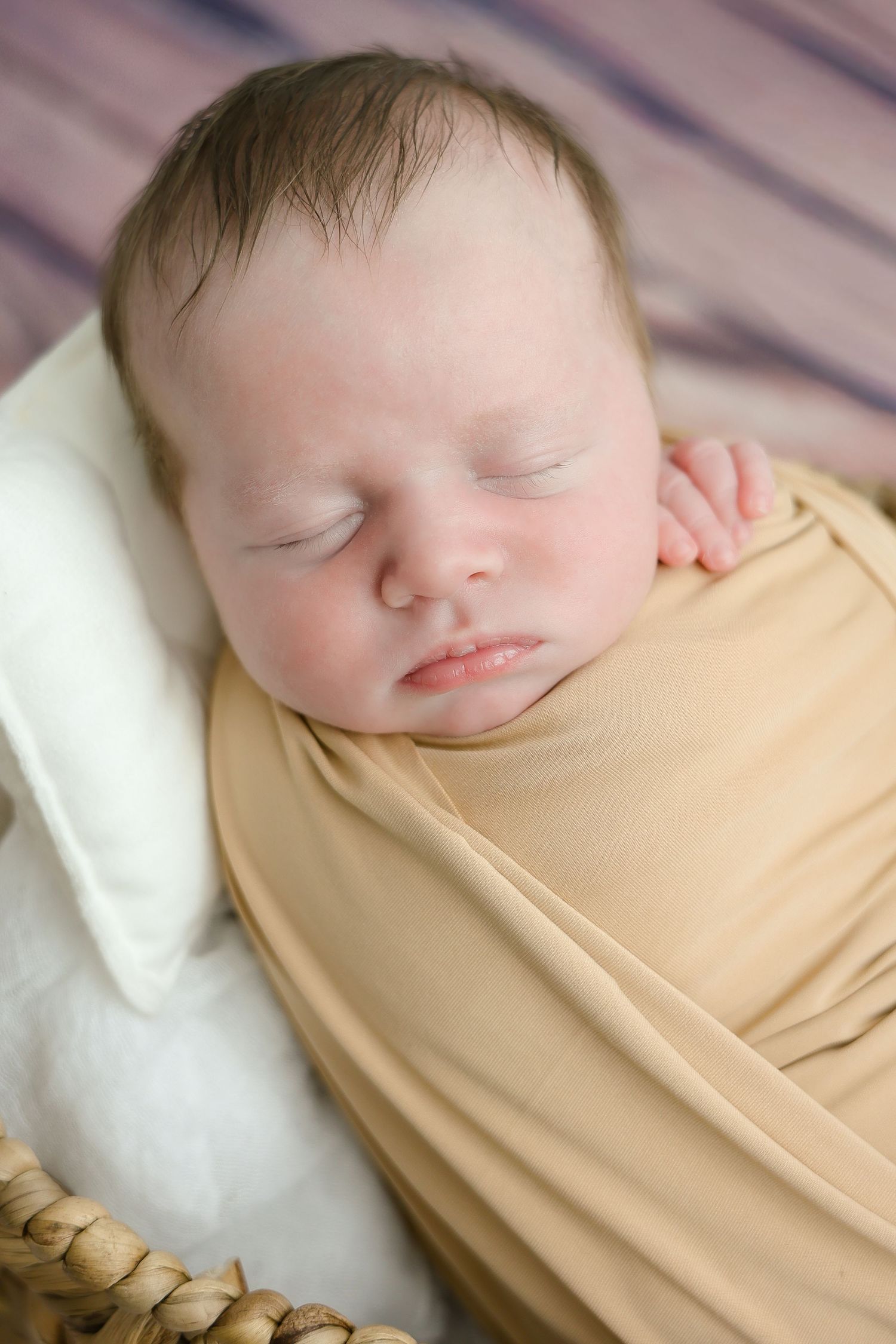 A sleeping baby wrapped in a soft beige swaddle blanket rests peacefully on white bedding.
