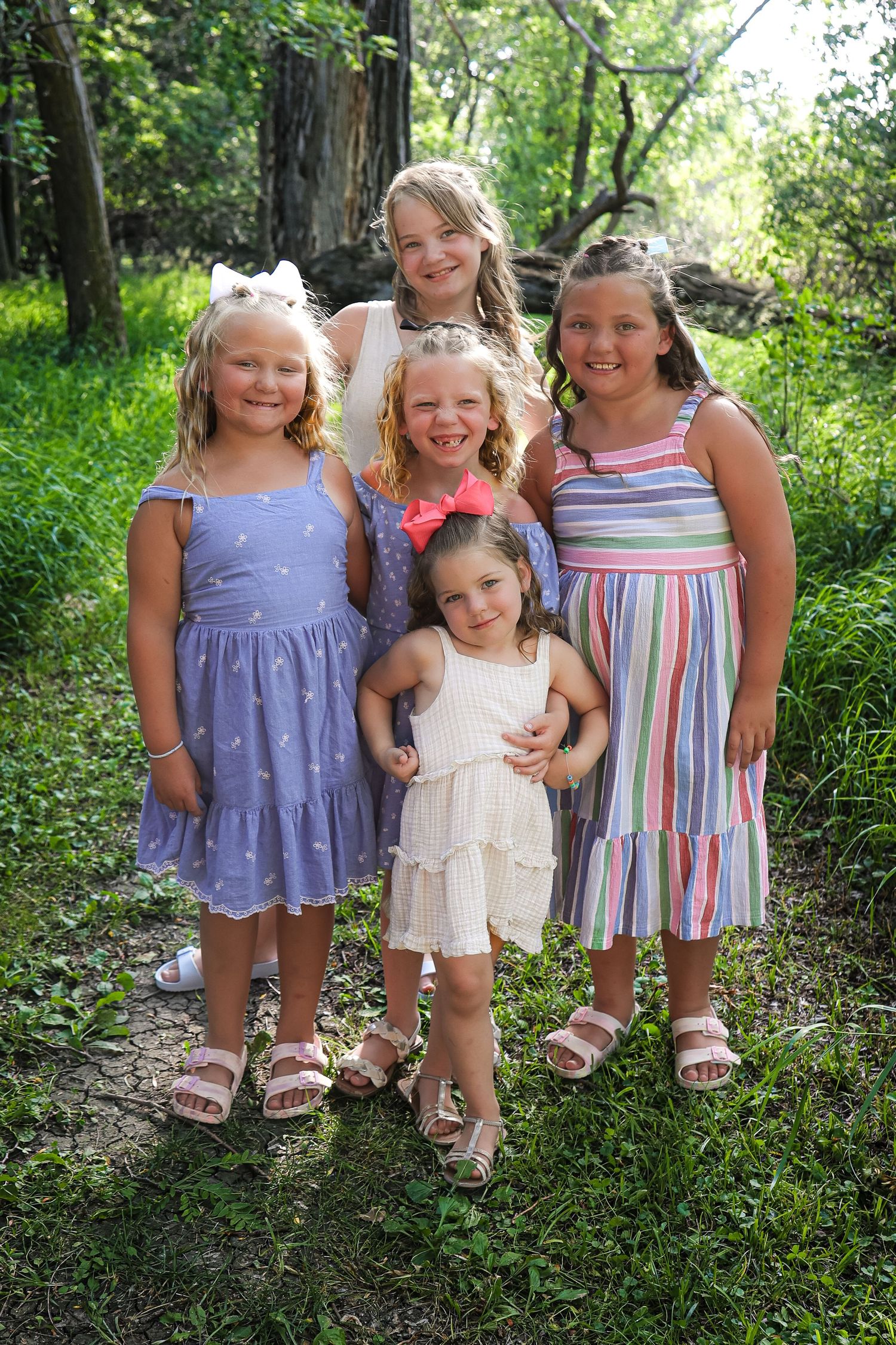 Children in summer dresses pose together on a woodland path surrounded by greenery.