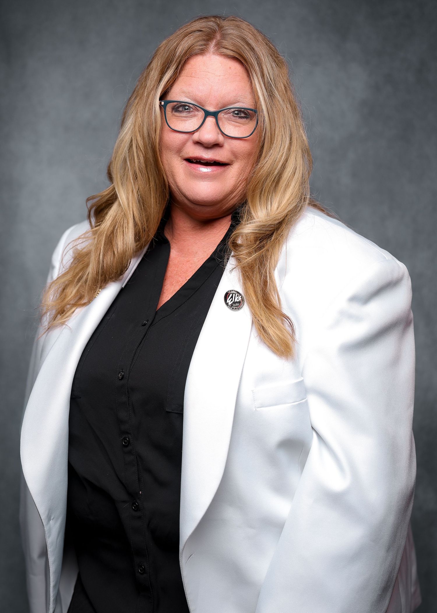 Woman with glasses in white blazer and black top wearing a pin smiling for professional headshot photo.