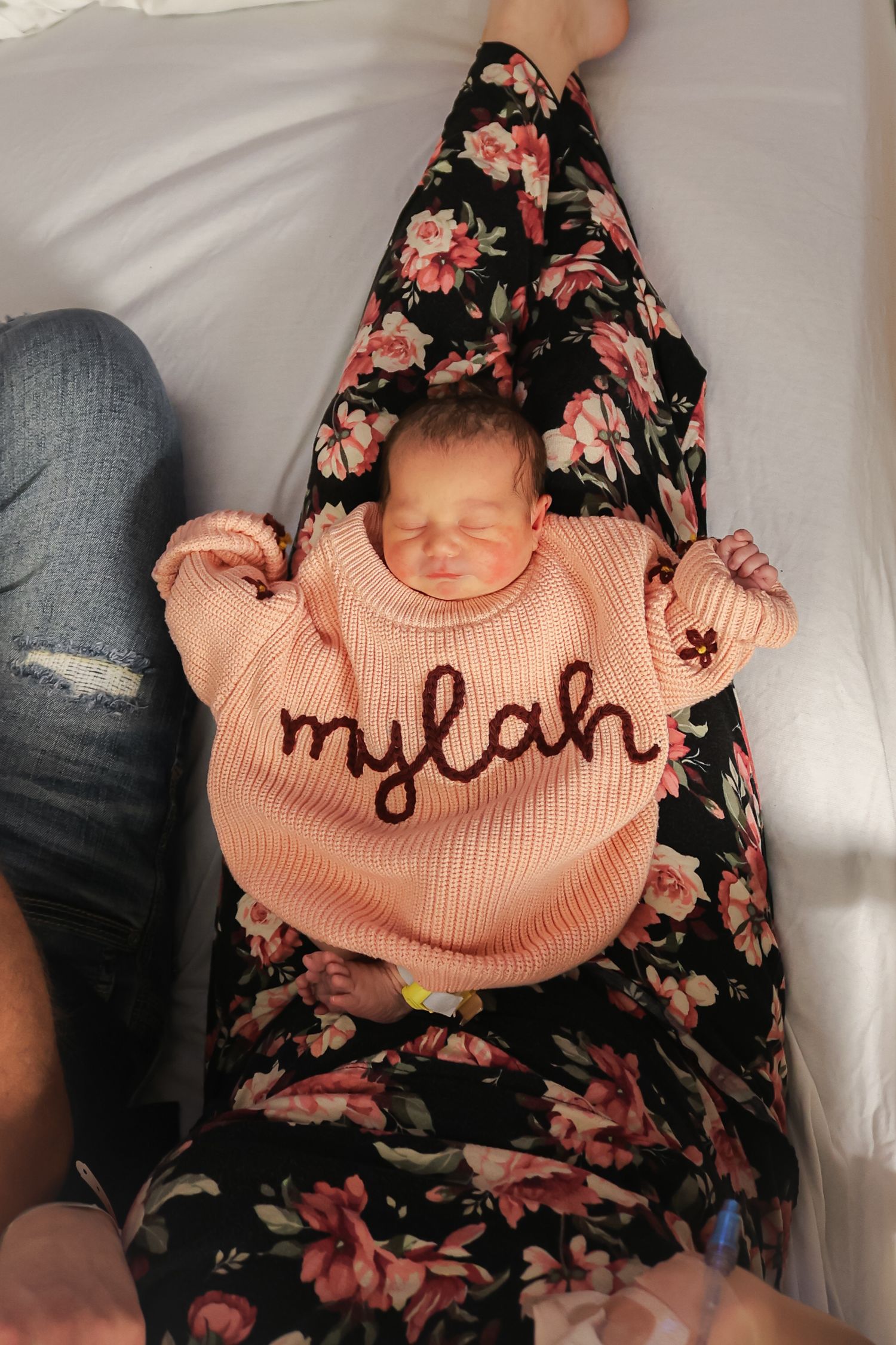 Newborn wearing personalized peach sweater lying on white hospital bedding.