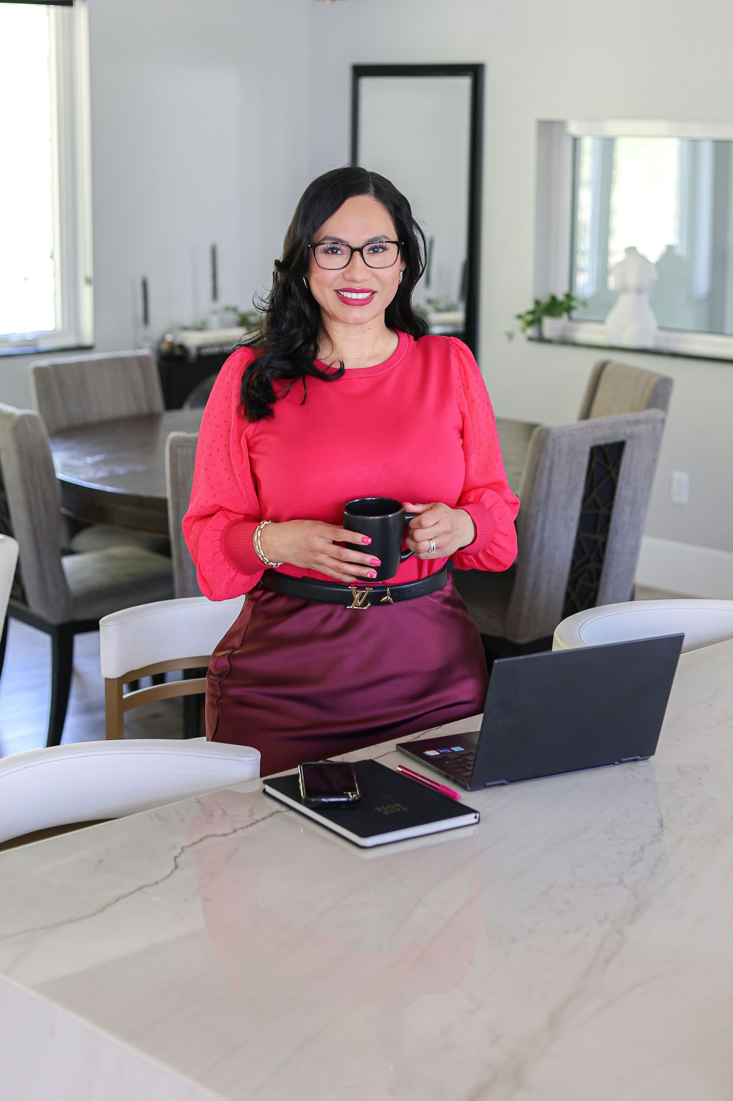 A professional woman in a red blouse and burgundy skirt holds a coffee mug while leaning against a marble desk with a laptop.