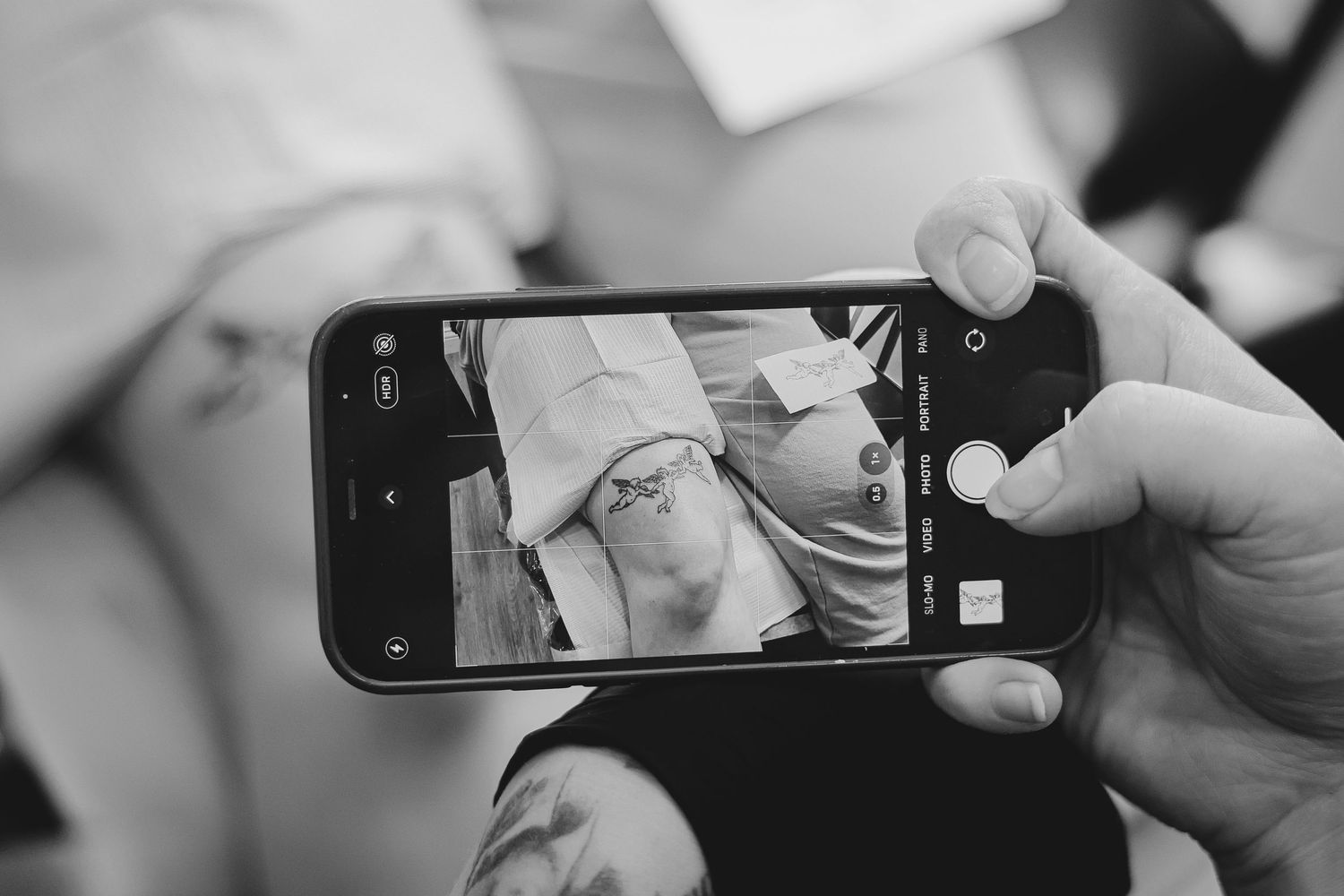 Black and white photo of hands holding a smartphone photographing a tattoo on someones skin.