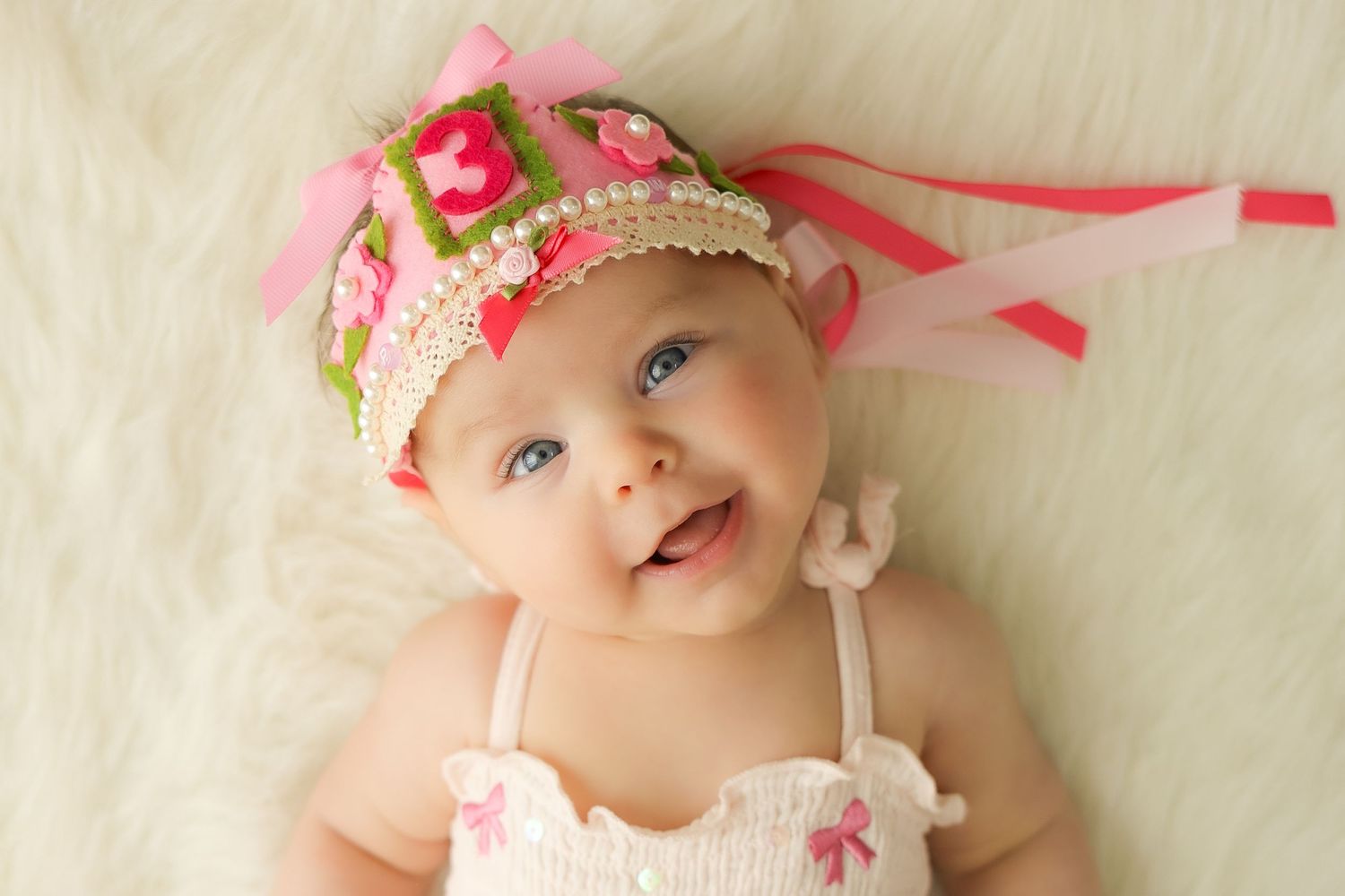 Smiling baby wearing a colorful pink and green crocheted headband with ribbons, lying on a white fluffy blanket.